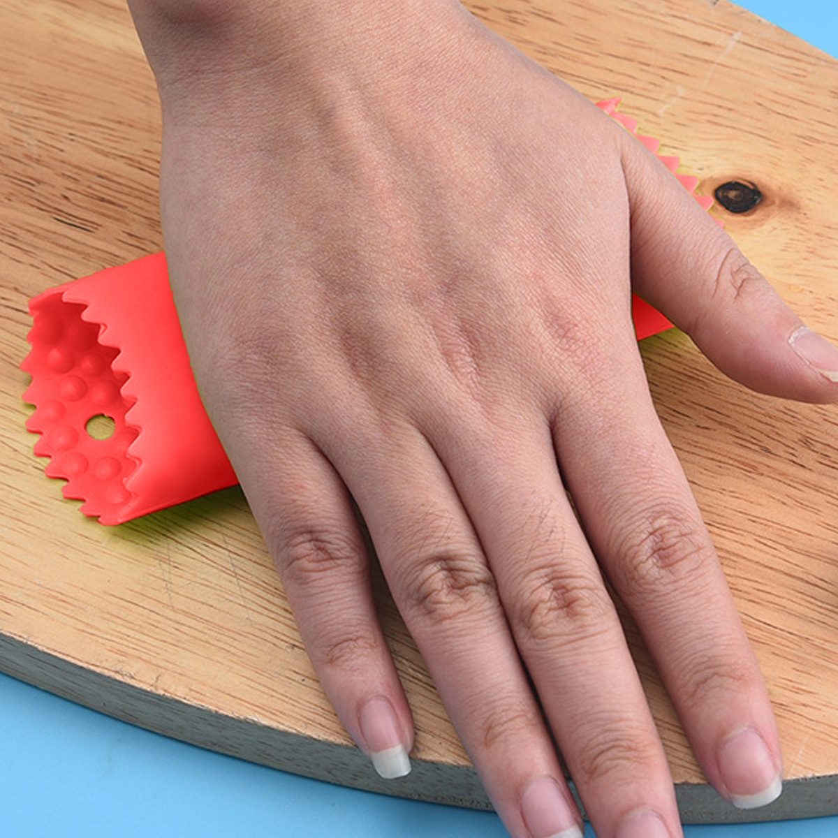 Eine Hand liegt auf einer Holzoberfläche und hält ein rotes Küchenwerkzeug. Das Werkzeug hat eine gezackte Kante und liegt neben der Hand.