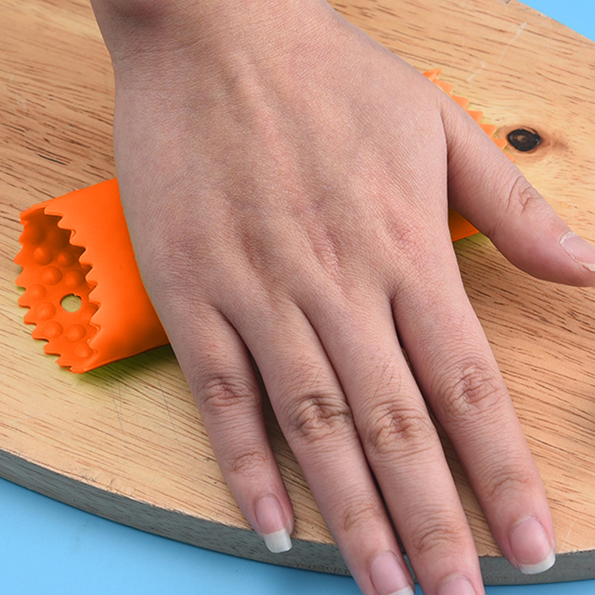 Eine Hand liegt auf einem Holzbrett und hält einen orangen Gemüseschäler. Der Schäler ist röhrenförmig mit gezacktem Rand. Die Nägel sind gepflegt.