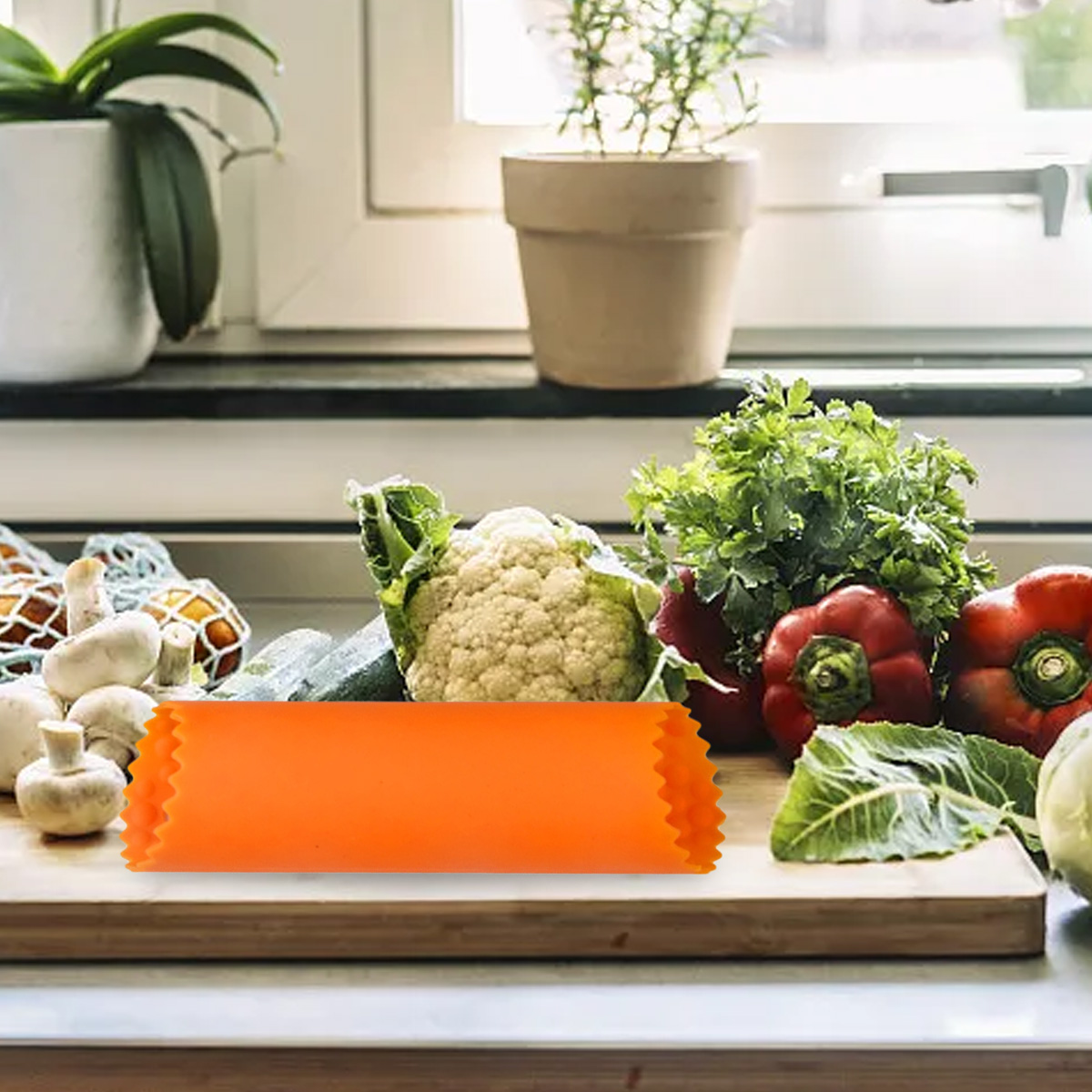 Ein orangefarbener Gemüseschäler liegt auf einem Holzbrett mit Gemüse wie Blumenkohl, roten Paprika und Grünzeug. Pflanzen sind im Hintergrund.
