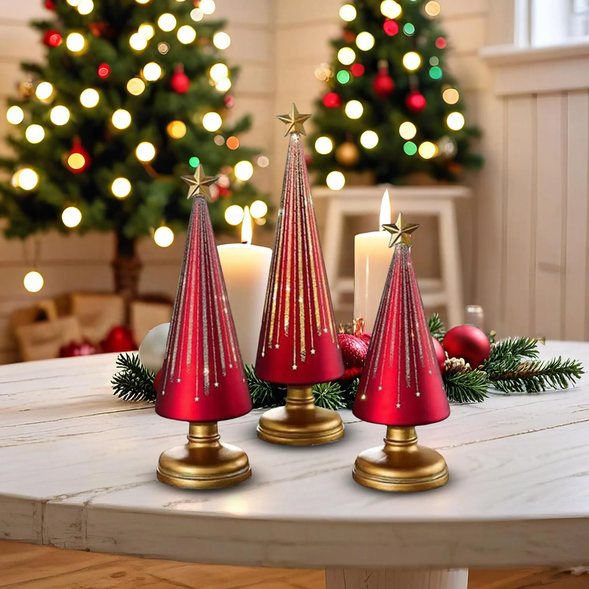 Drei rote Weihnachtsbaumdekorationen mit goldenen Sternen auf einem weißen Tisch. Zwei beleuchtete Weihnachtsbäume im Hintergrund.