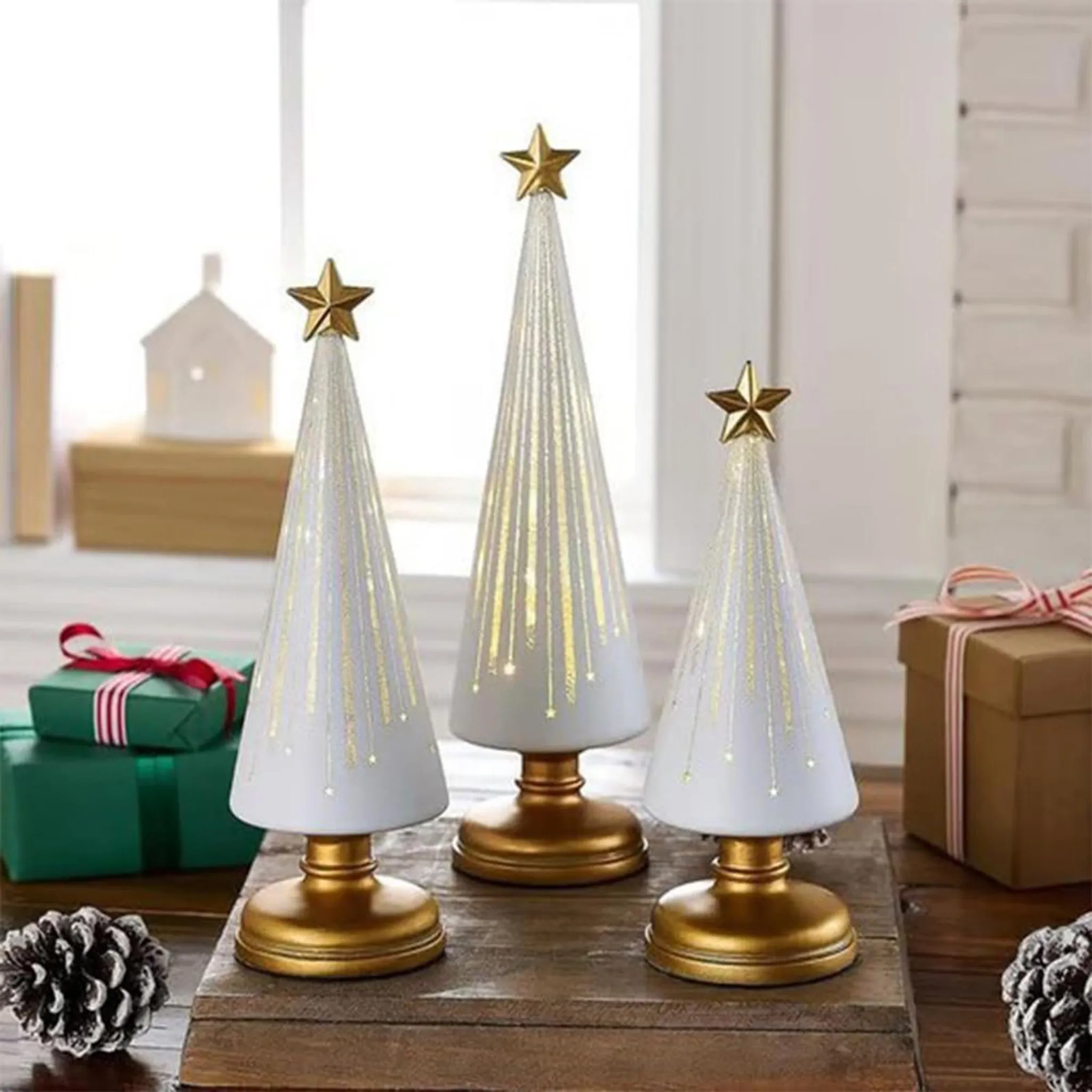 Drei weiße Weihnachtsbäume mit goldenen Sternen auf einer Holzoberfläche mit Geschenken und Tannenzapfen.