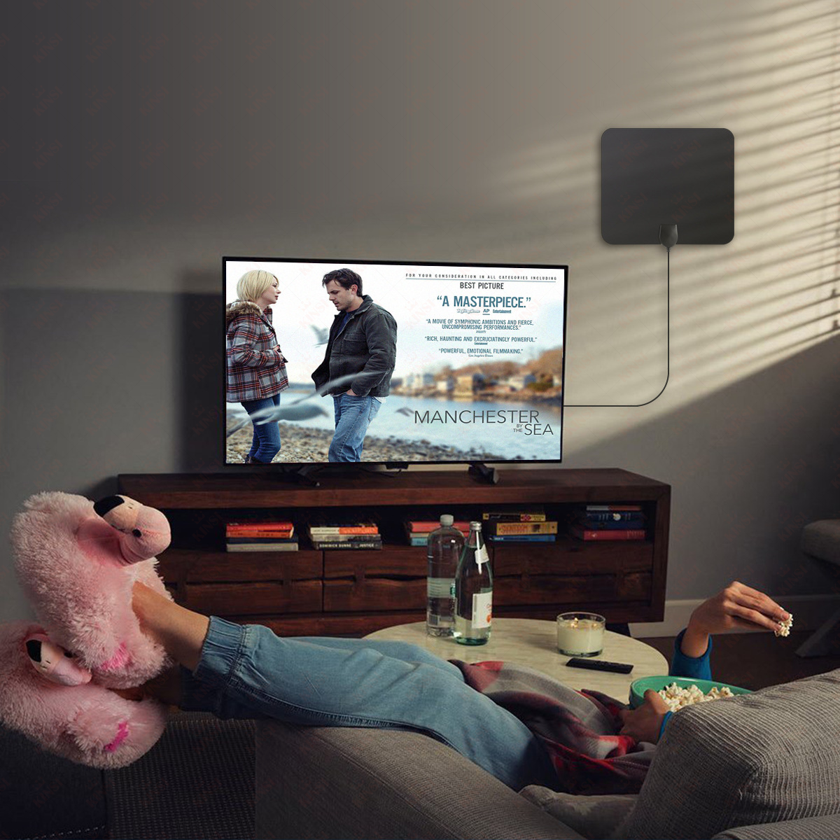Person entspannt auf einer Couch, schaut fern mit rosa Flamingo-Hausschuhen und isst Popcorn. Der Fernseher zeigt 'Manchester by the Sea'.