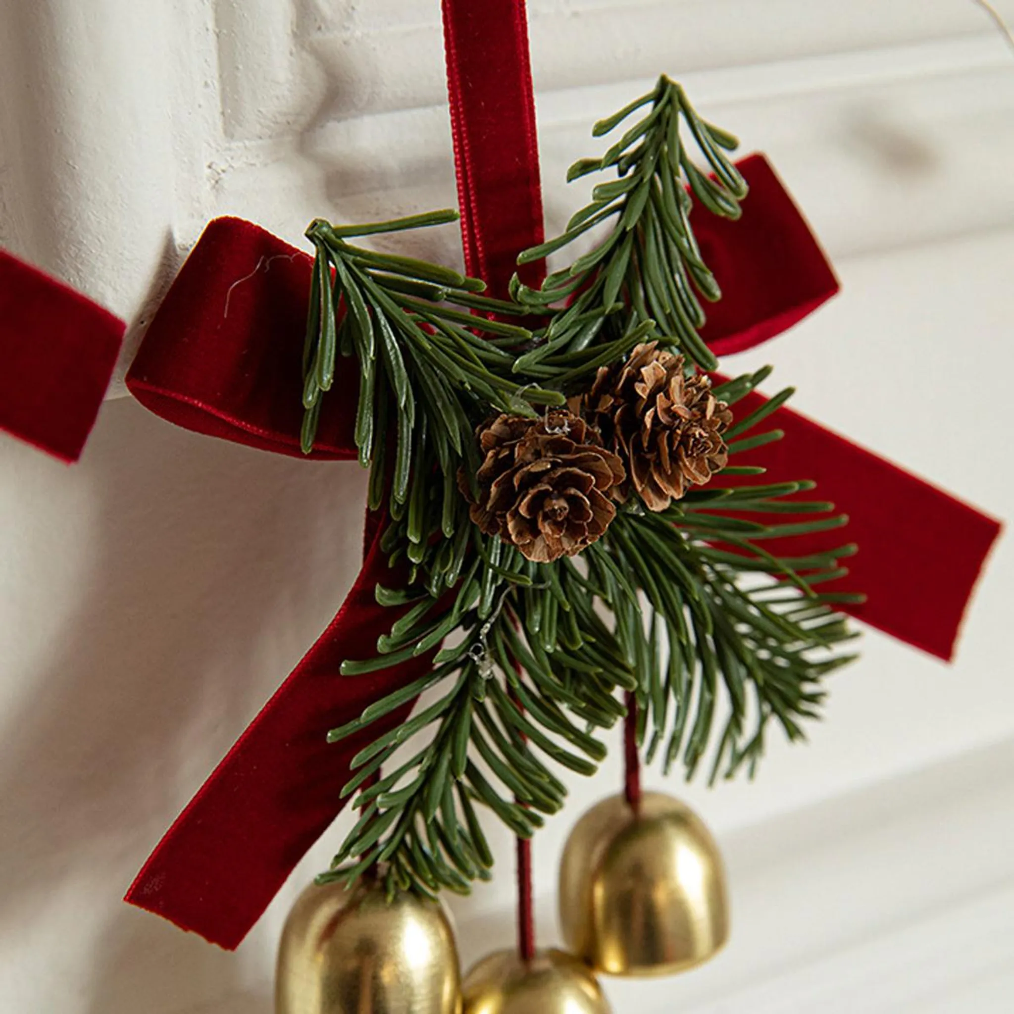 Eine Weihnachtsdekoration mit rotem Band, Tannenzapfen, grünen Nadeln und goldenen Glocken vor weißem Hintergrund.