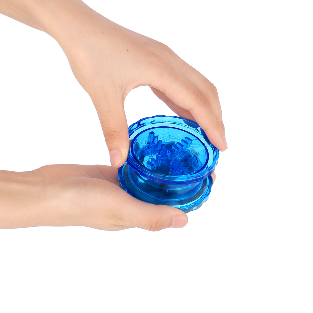 Hände halten einen transparenten, blauen Grinder. Der Grinder besteht aus zwei kreisförmigen Teilen. Der Hintergrund ist weiß.