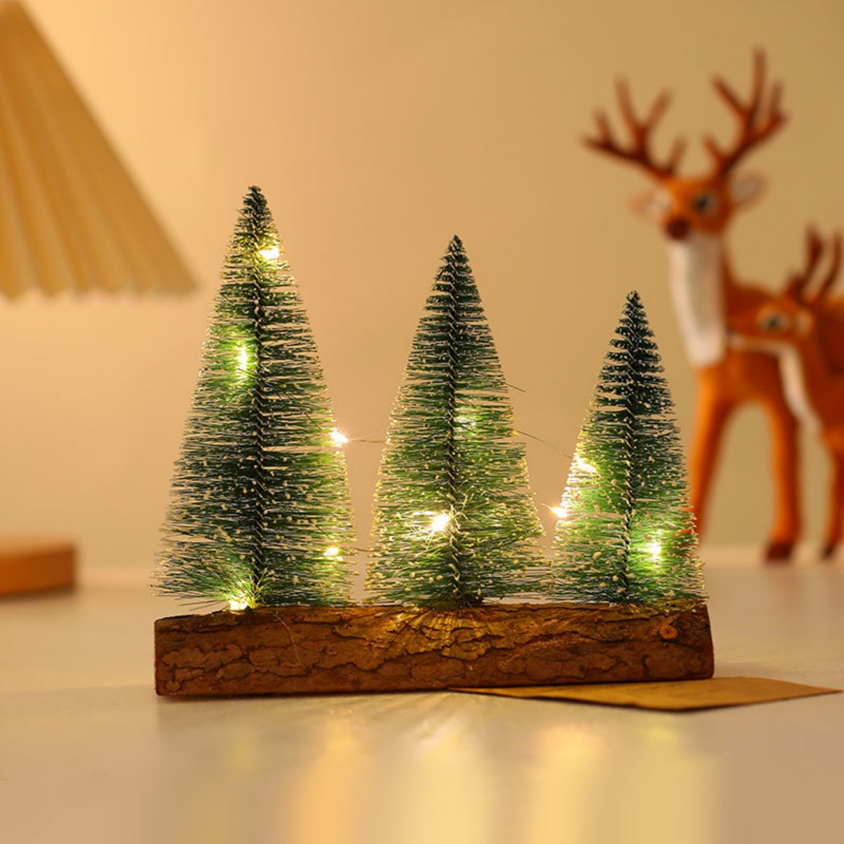 Drei Miniatur-Weihnachtsbäume mit Lichtern auf einem Holzsockel. Eine Rentierfigur verschwommen im Hintergrund, festliche Szene.