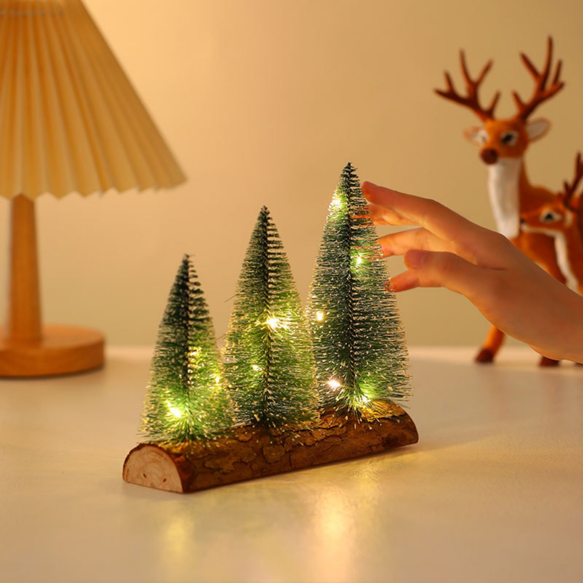 Drei Miniatur-Weihnachtsbäume mit Lichtern auf einem Holzsockel. Eine Hand greift danach. Im Hintergrund eine Hirschfigur.