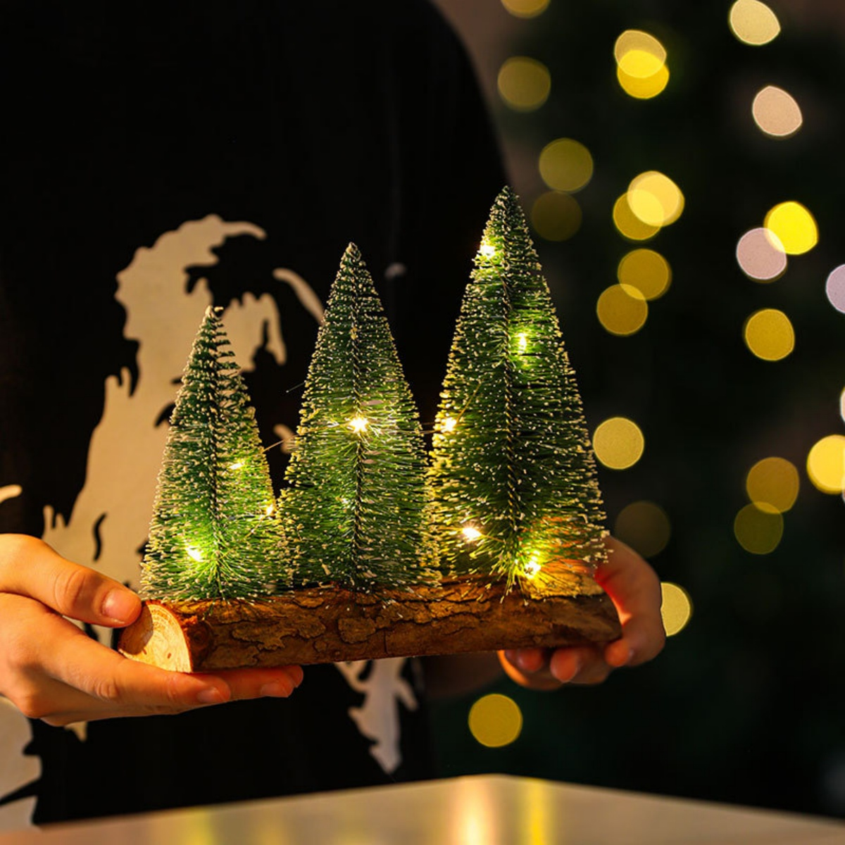 Person hält eine Holzbasis mit drei kleinen, beleuchteten Weihnachtsbäumen. Der Hintergrund hat Bokeh-Lichter und einen schwarzen Hintergrund.