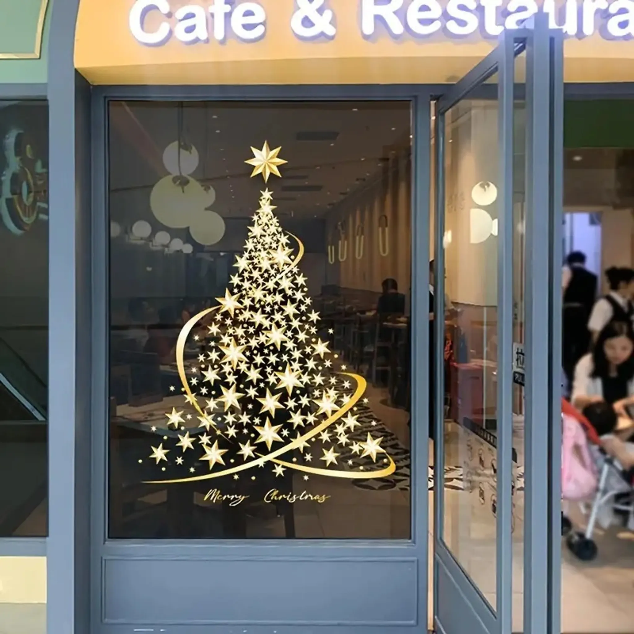Ein Café-Eingang mit Glastür. Auf dem Glas befindet sich ein goldener Weihnachtsbaum. Menschen sind drinnen und draußen sichtbar.