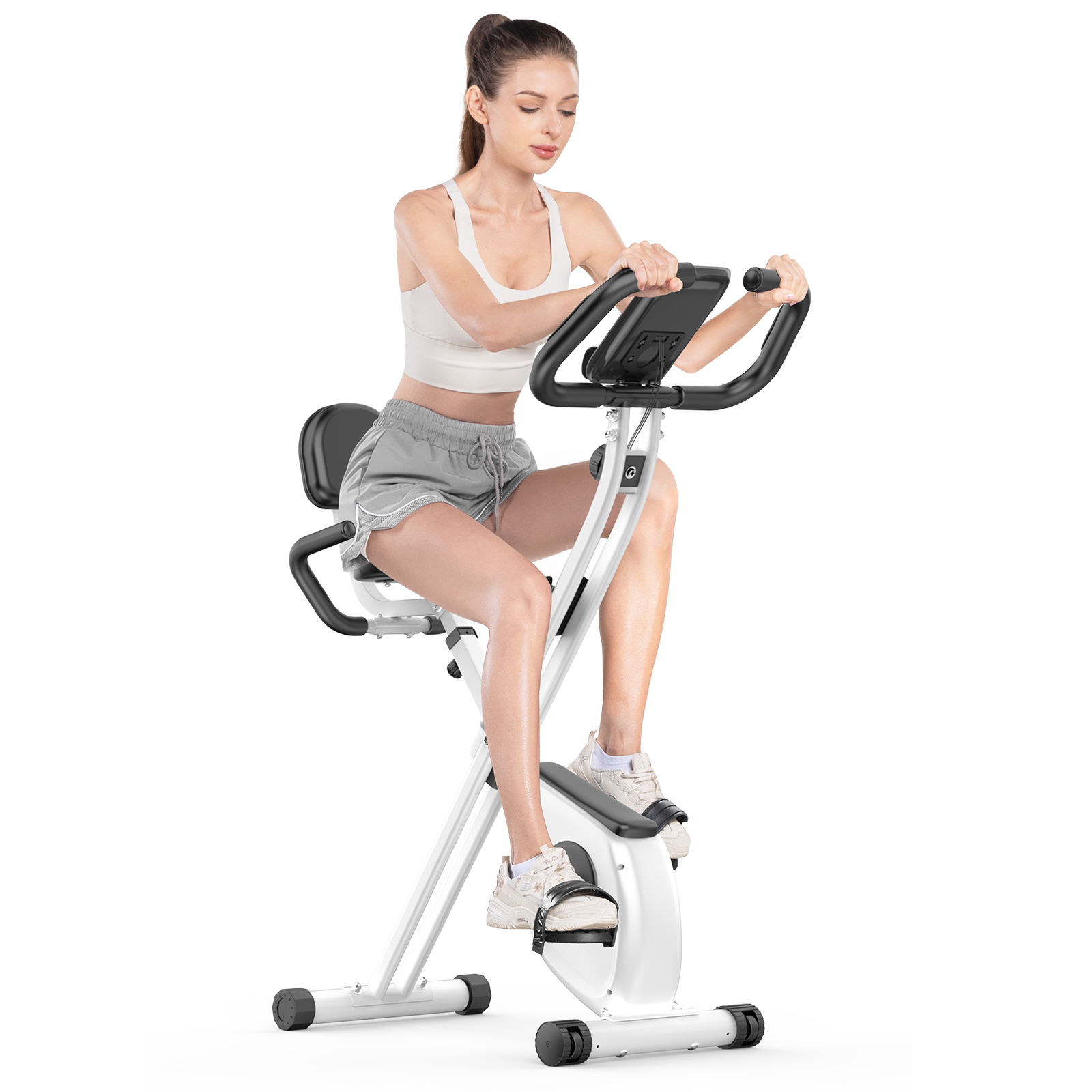 Kobieta ćwiczy na rowerze stacjonarnym. Ma na sobie biały top, szare szorty i białe trampki. Rower jest biało-czarny.