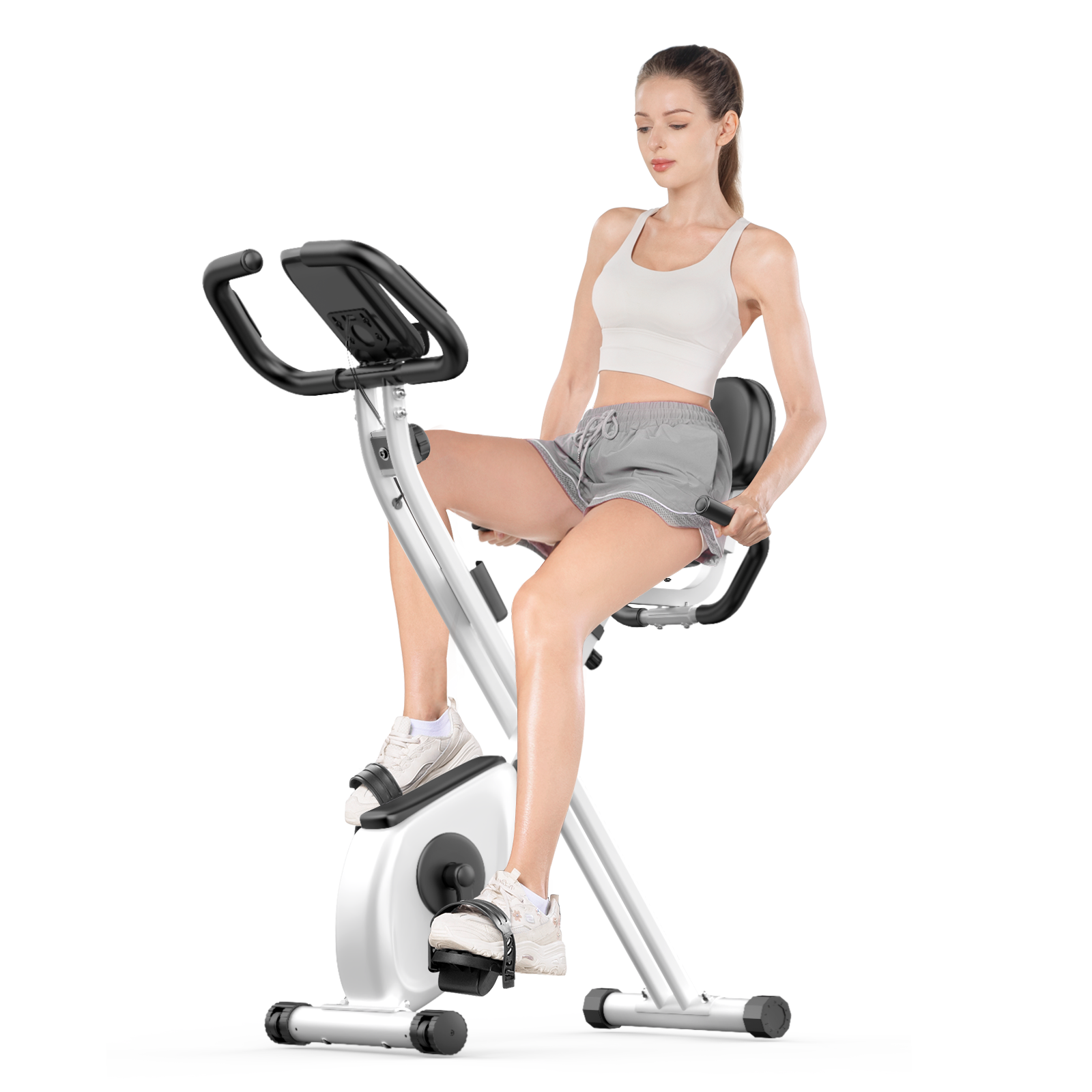Kobieta na rowerze stacjonarnym. Ma na sobie biały top, szare szorty i białe trampki. Rower jest biało-czarny.