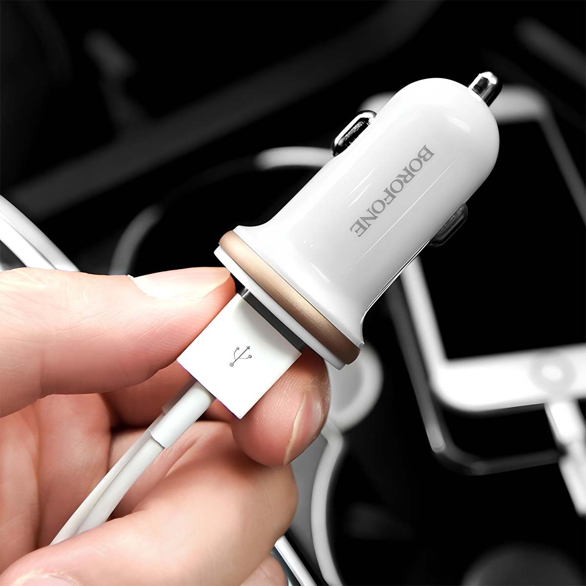 Un caricabatterie per auto Borofone bianco e oro è tenuto, con un cavo USB collegato. Lo sfondo è sfocato, mostrando interni auto.