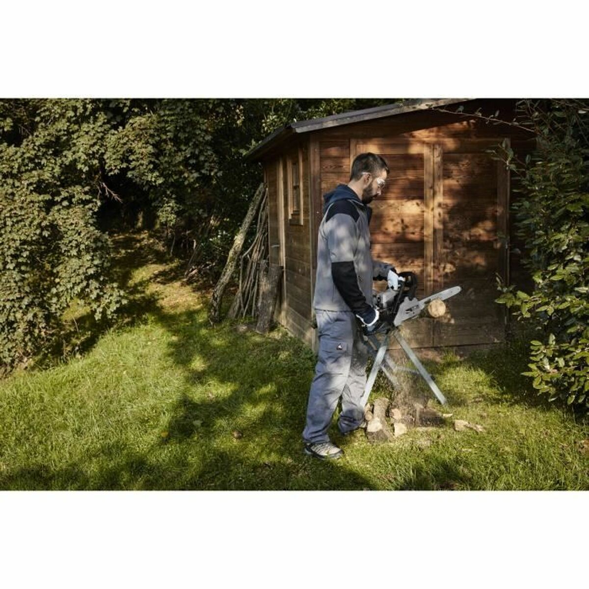 Ein Mann benutzt eine Kettensäge an einem Baumstamm. Im Hintergrund befindet sich eine Holzhütte. Grünes Gras und Bäume umgeben die Szene.