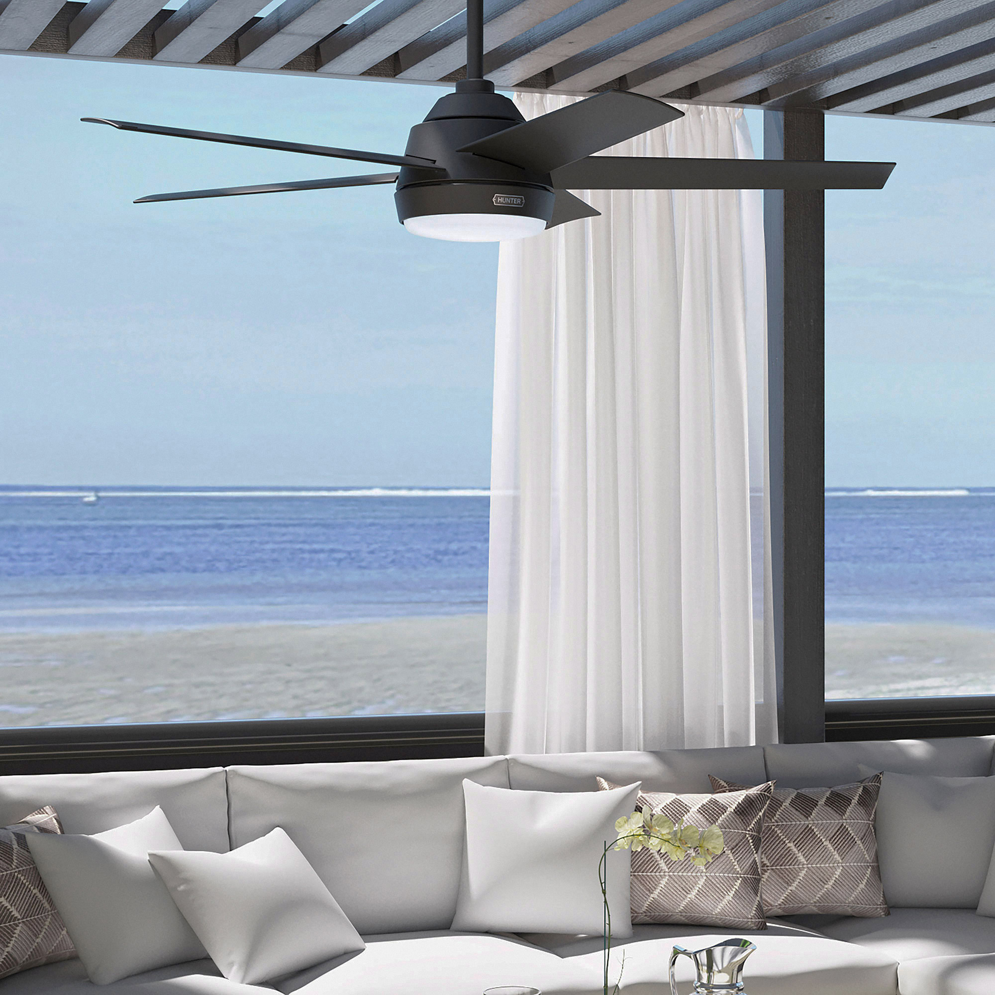 Ein Deckenventilator über einem weißen Sofa mit Kissen. Durch das Fenster sieht man Strand und Meer.