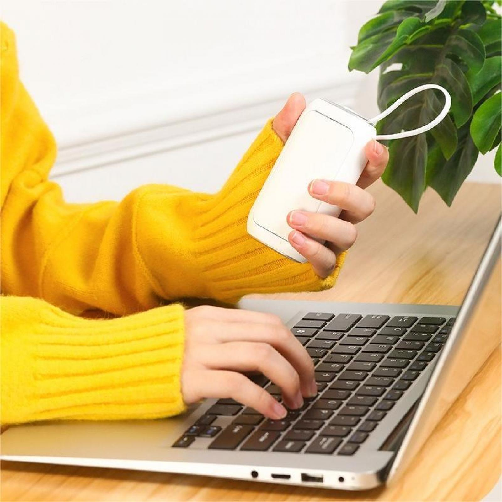 Eine Person in einem gelben Pullover hält ein weißes Gerät und tippt auf einem Laptop. Grüne Pflanze im Hintergrund.