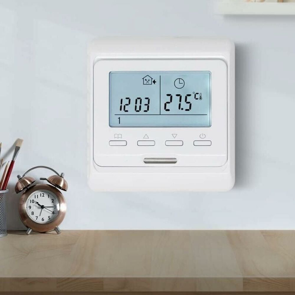 Ein weißer Thermostat an einer weißen Wand zeigt Zeit und Temperatur. Eine Uhr und Bleistifte stehen auf einer Holzoberfläche.