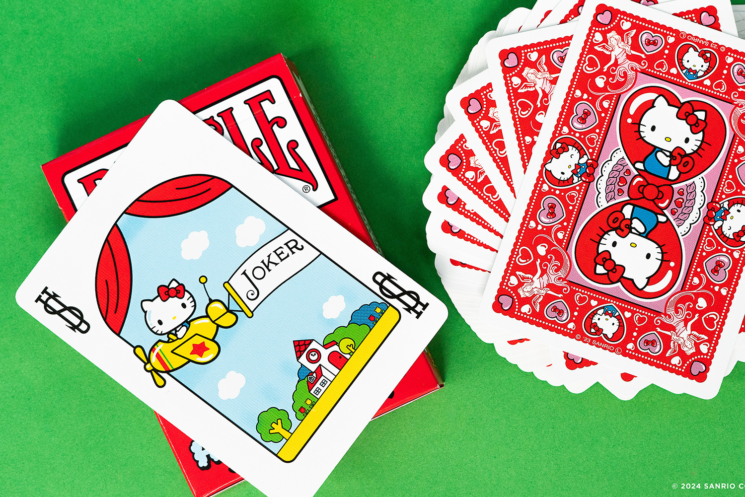 Hello Kitty Spielkarten. Joker-Karte mit Kitty im Flugzeug. Rote Kartenrücken mit Kitty und Herzen. Grüner Hintergrund.