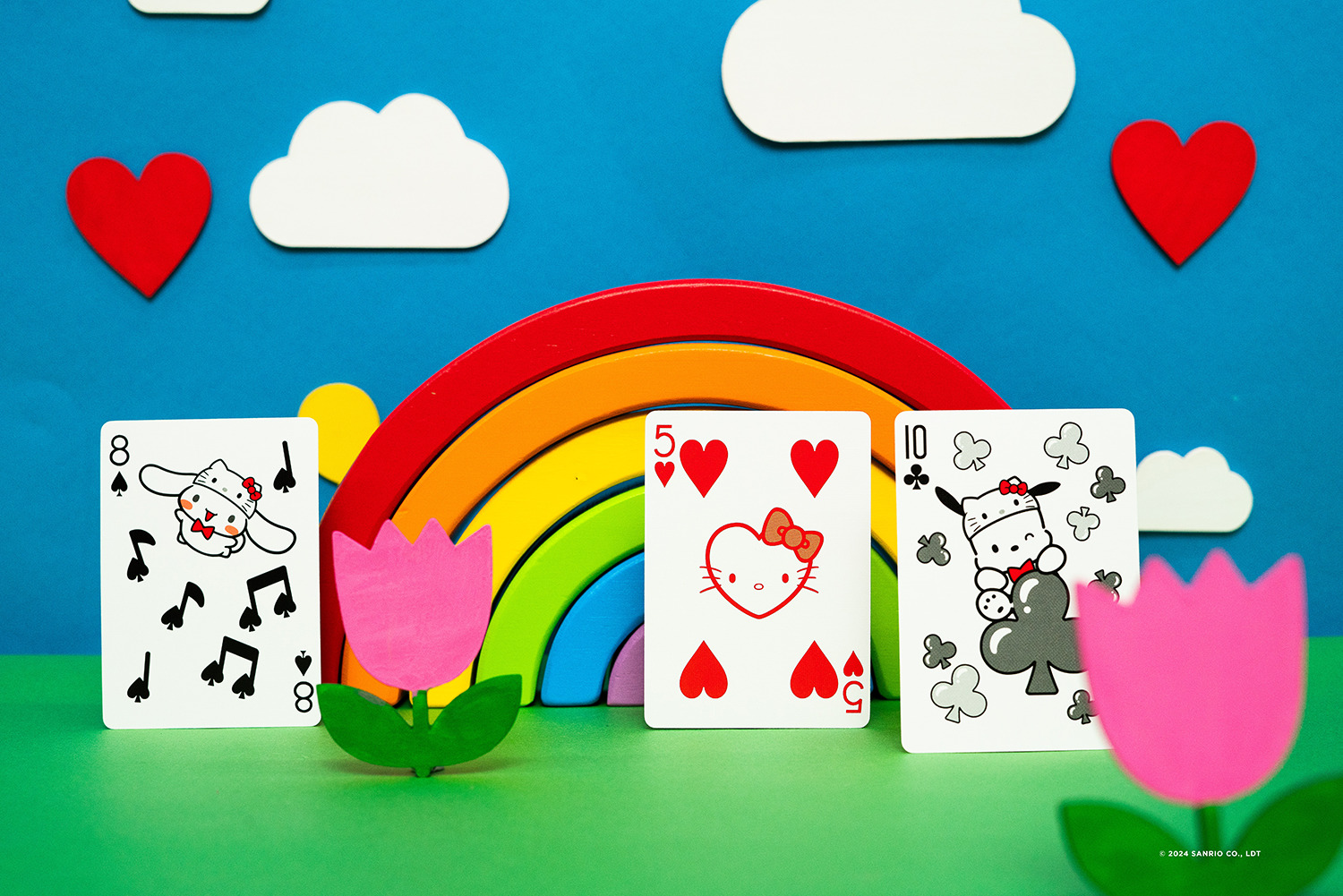 Karten mit Zeichentrickfiguren, Regenbogen, Herzen und Wolken auf blauem Grund. Rosa Tulpen im Vordergrund und grünes Gras.