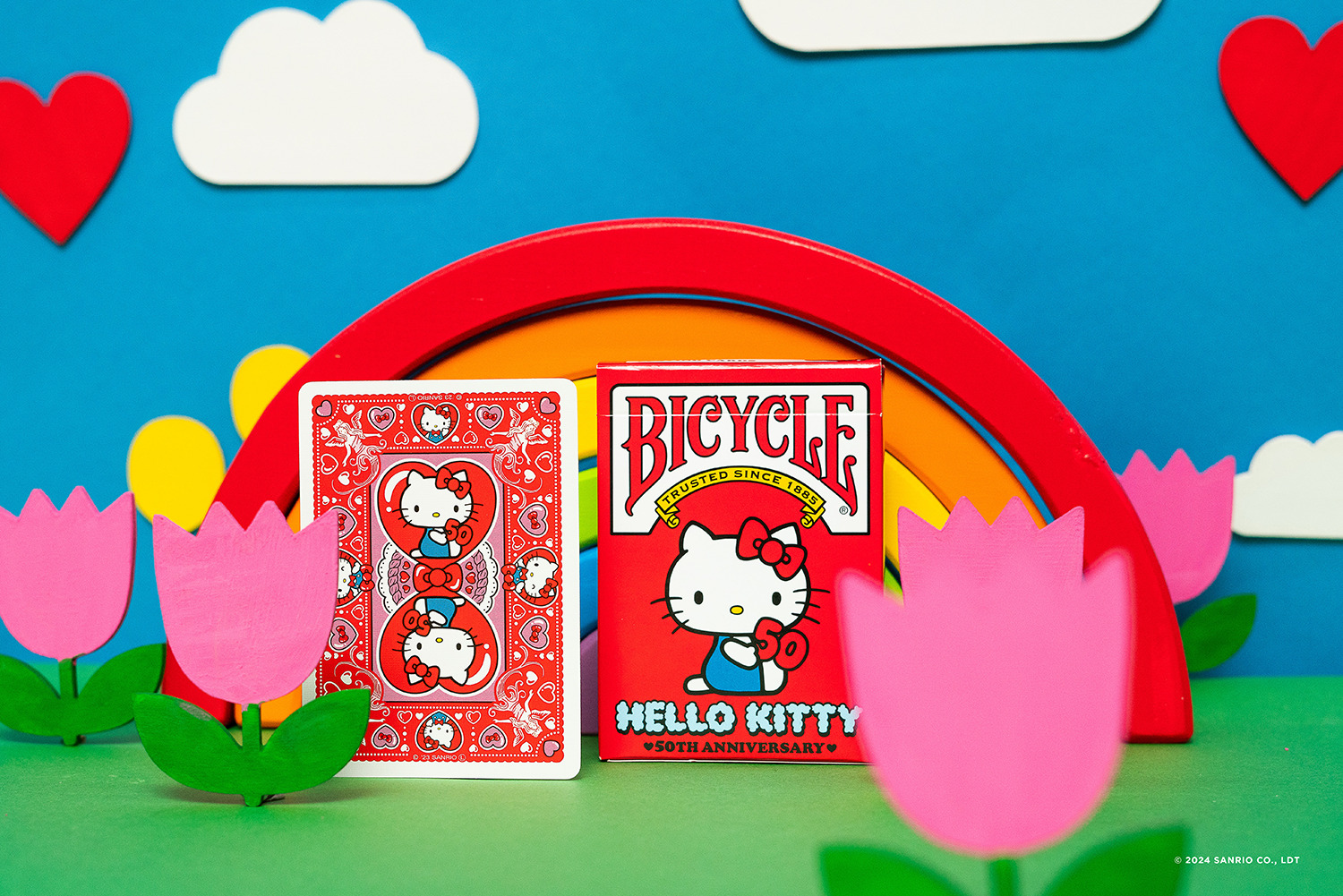 Ein Hello Kitty Bicycle Deck und eine Spielkarte sind mit einem Regenbogen, Wolken, Blumen und Herzen vor blauem Hintergrund abgebildet.