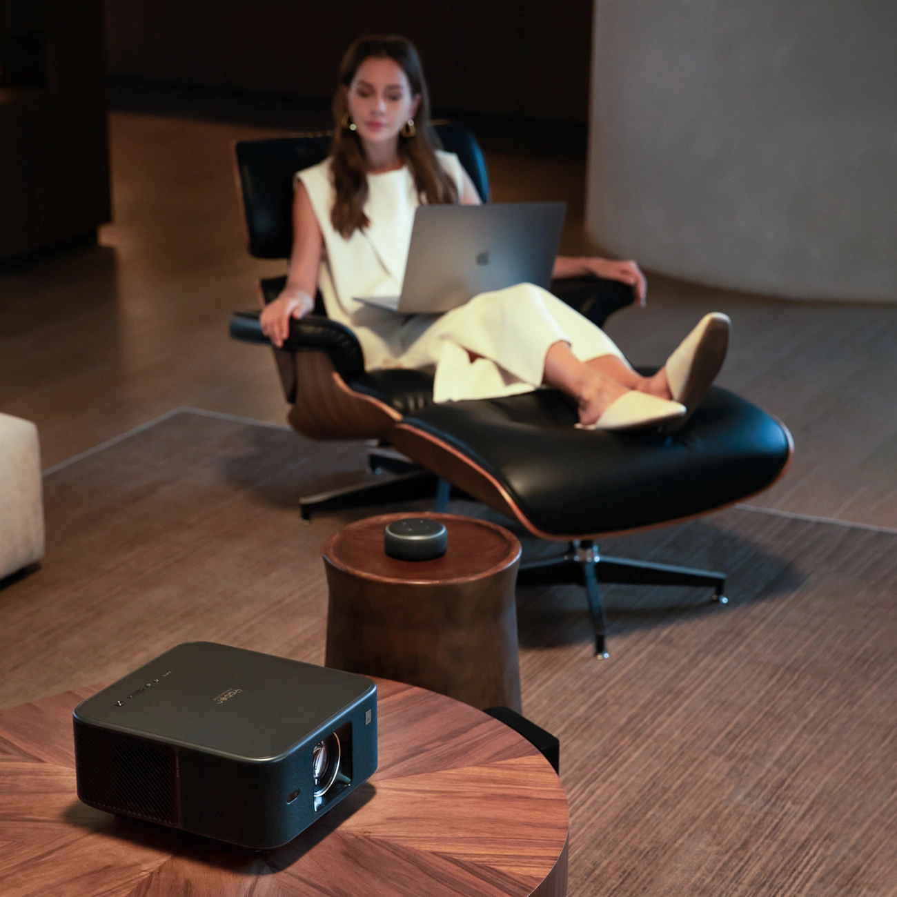 Kobieta z laptopem w fotelu. Projektor, głośnik i stół na pierwszym planie. Drewniana podłoga i neutralne ściany w tle.