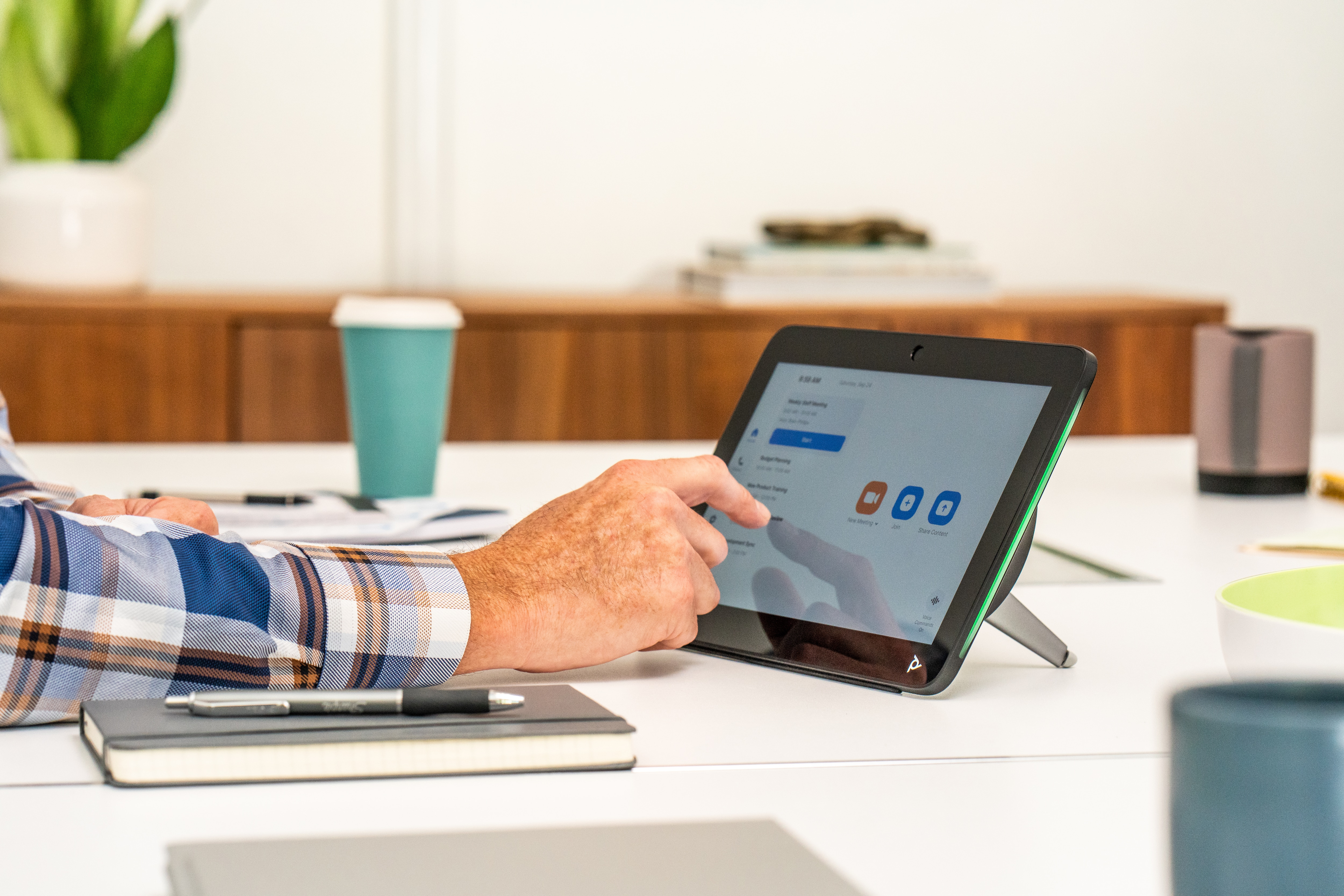 Eine Person benutzt ein Tablet für einen Videoanruf. Auf dem weißen Schreibtisch liegen Stift, Notizbuch und eine Tasse.