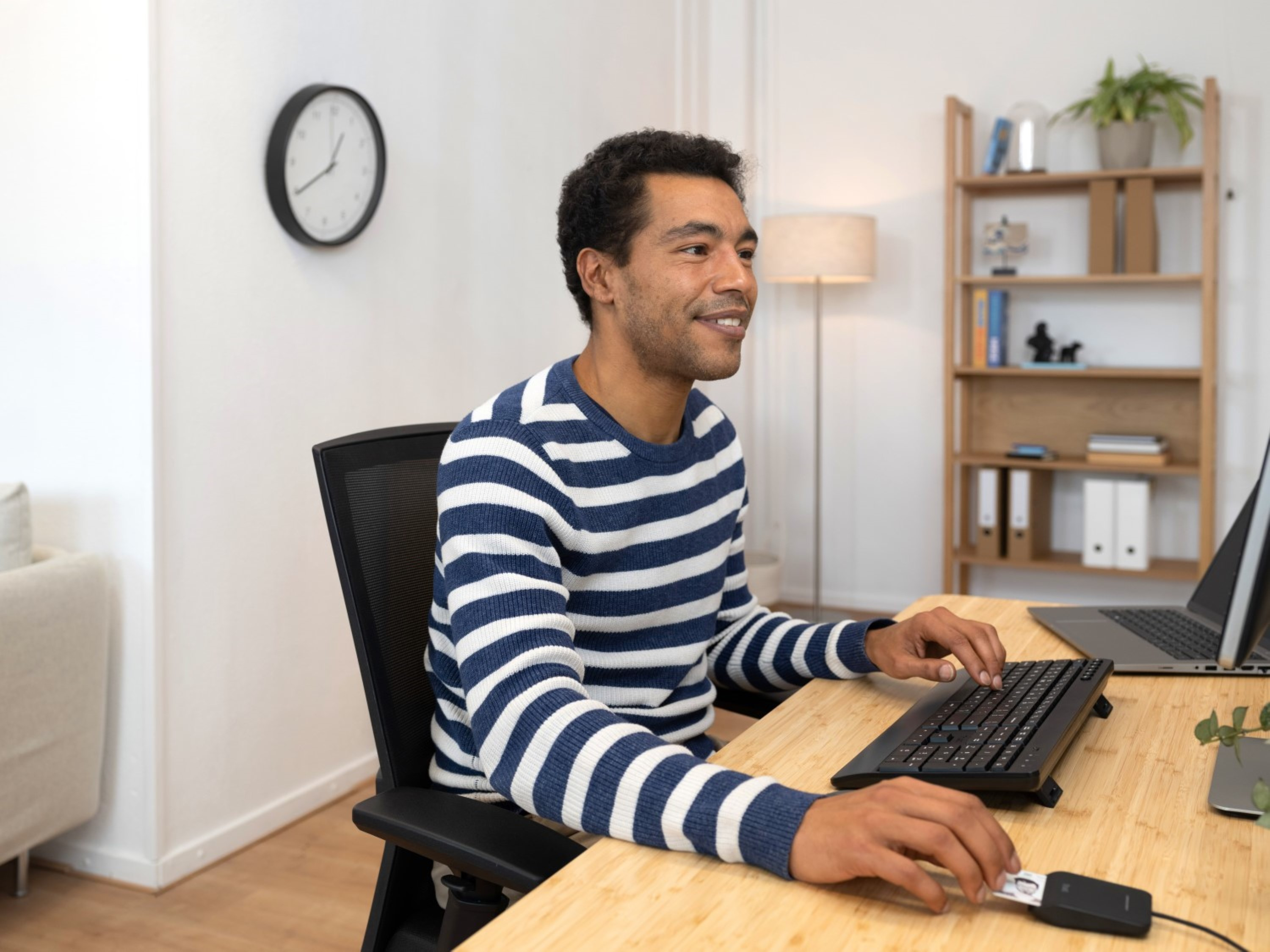 Ein Mann in gestreiftem Hemd arbeitet am Computer. Eine Uhr hängt an der Wand, dahinter ein Regal.