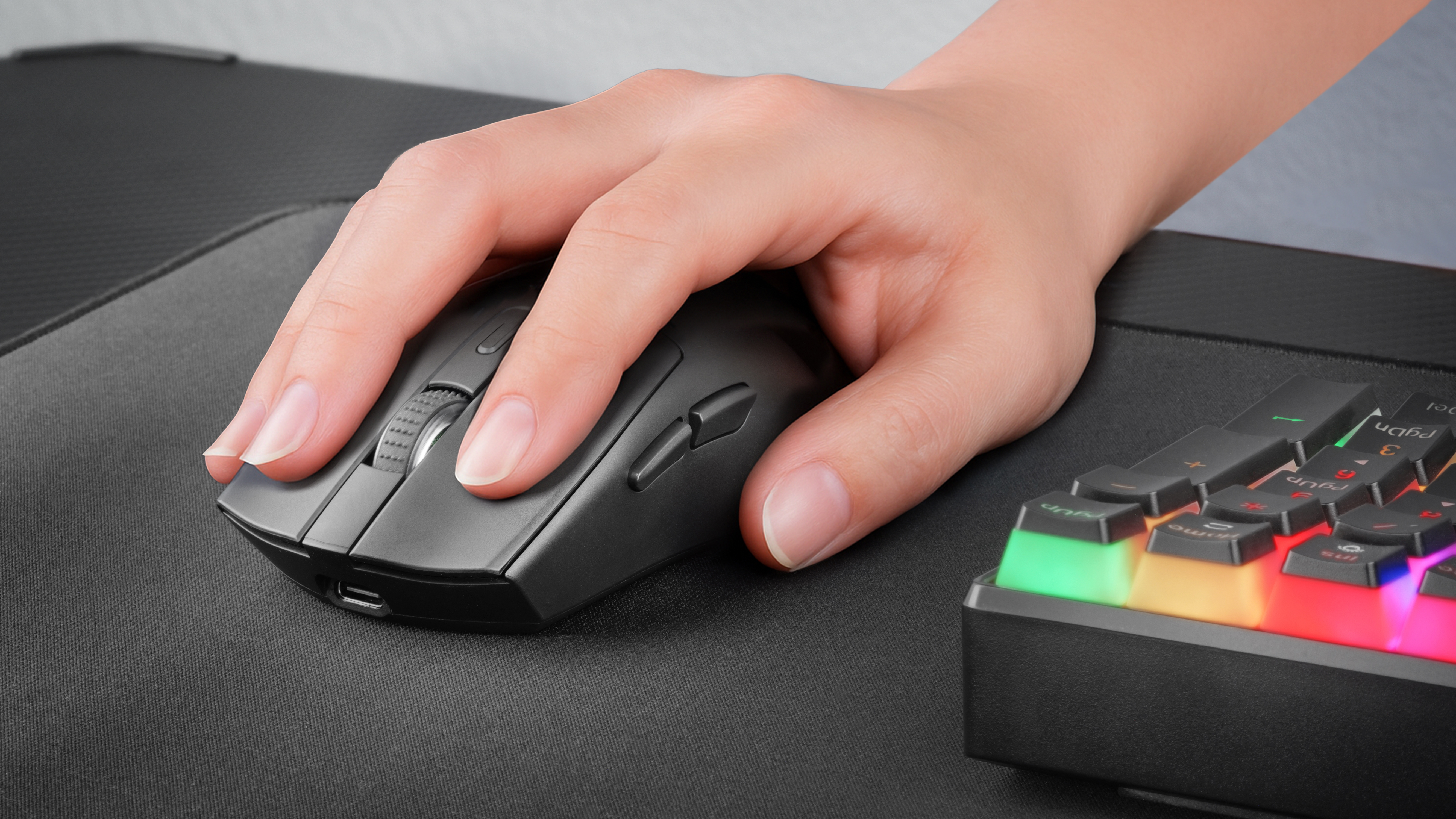 Dłoń spoczywa na szarej myszy na czarnej podkładce, obok podświetlanej klawiatury z kolorowymi klawiszami.