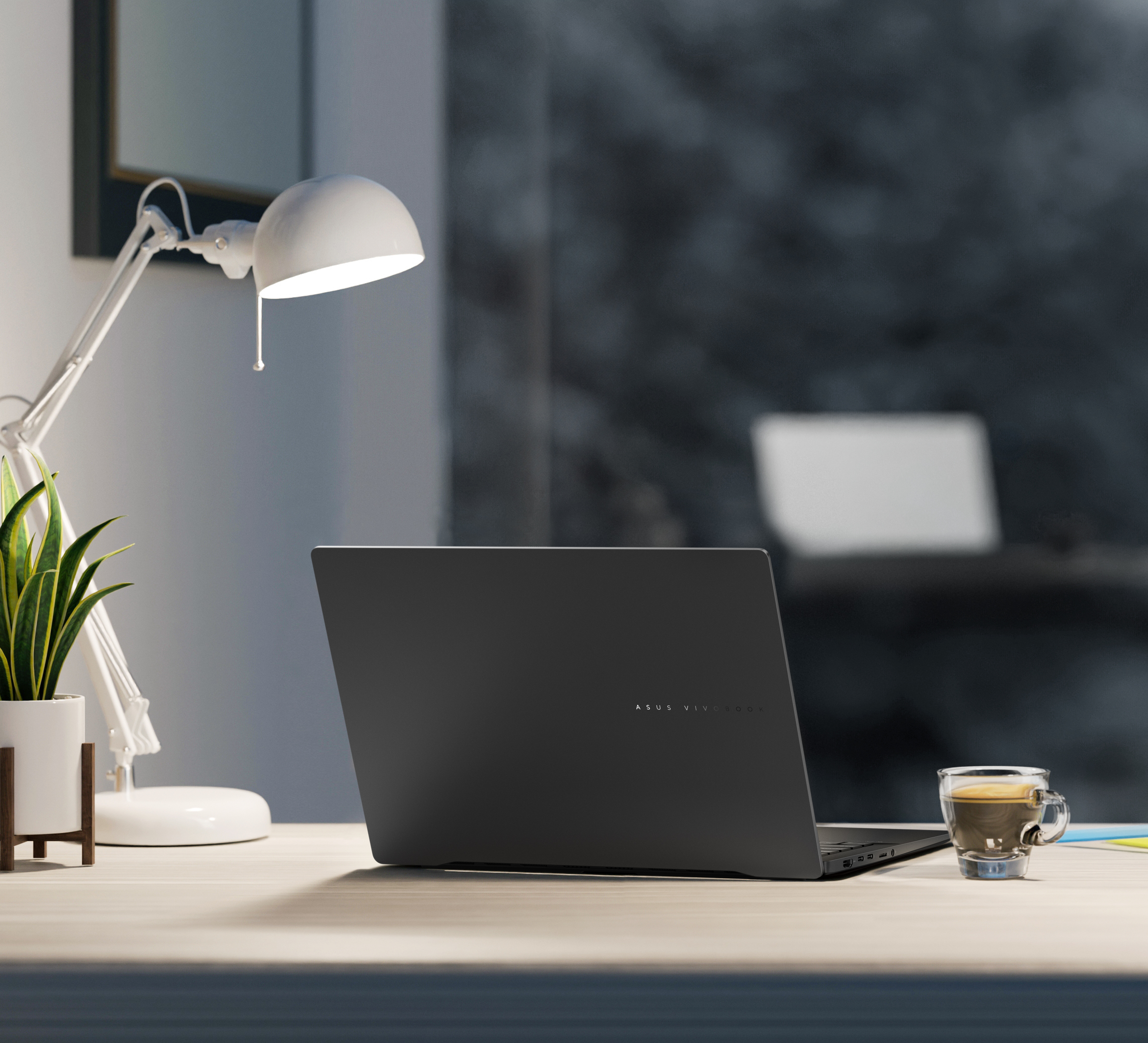 Laptop na biurku z lampą, rośliną i filiżanką kawy. Tło jest rozmyte.