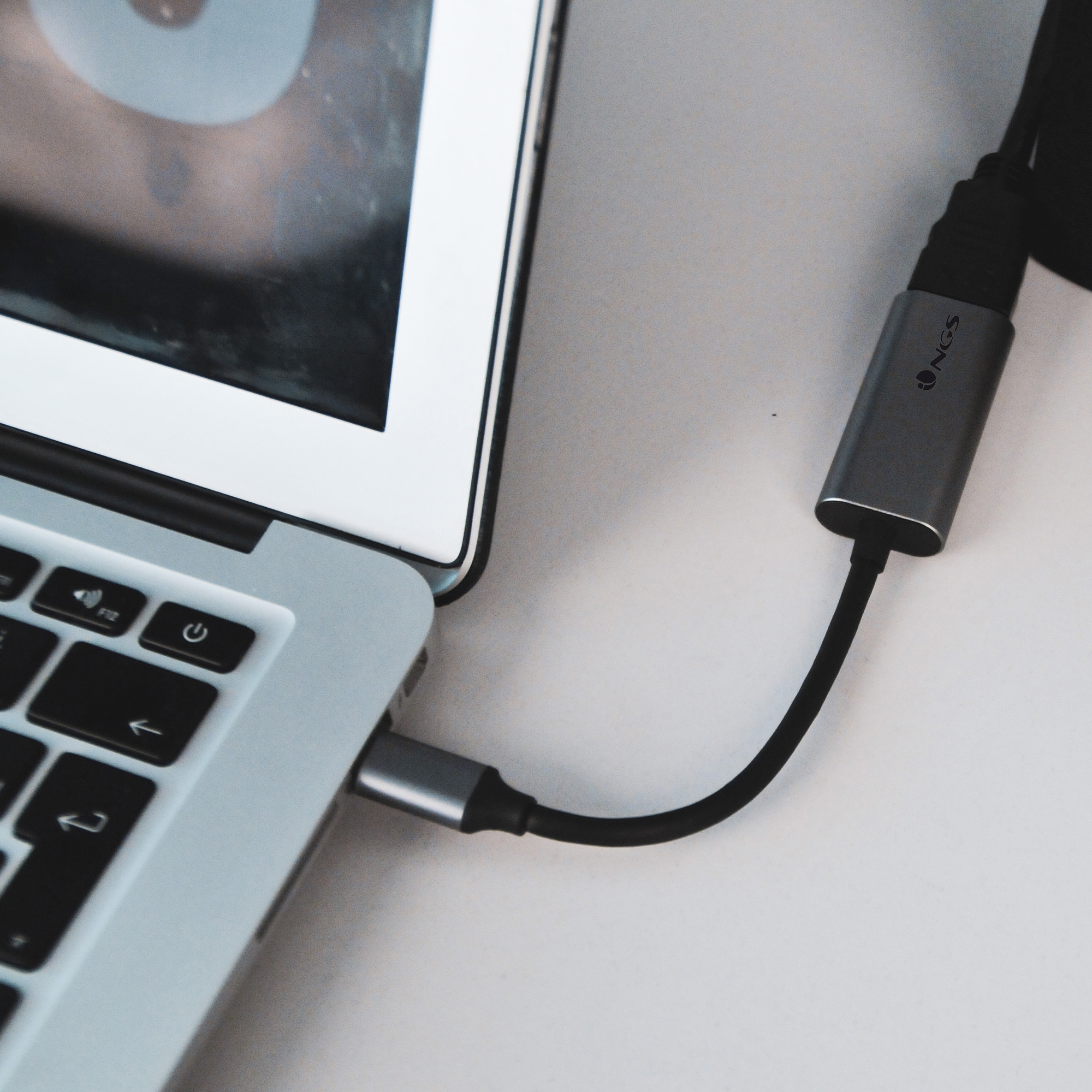 Ordenador portátil con un adaptador externo conectado. El cable y el adaptador son negros y plateados. El portátil está sobre una superficie blanca.