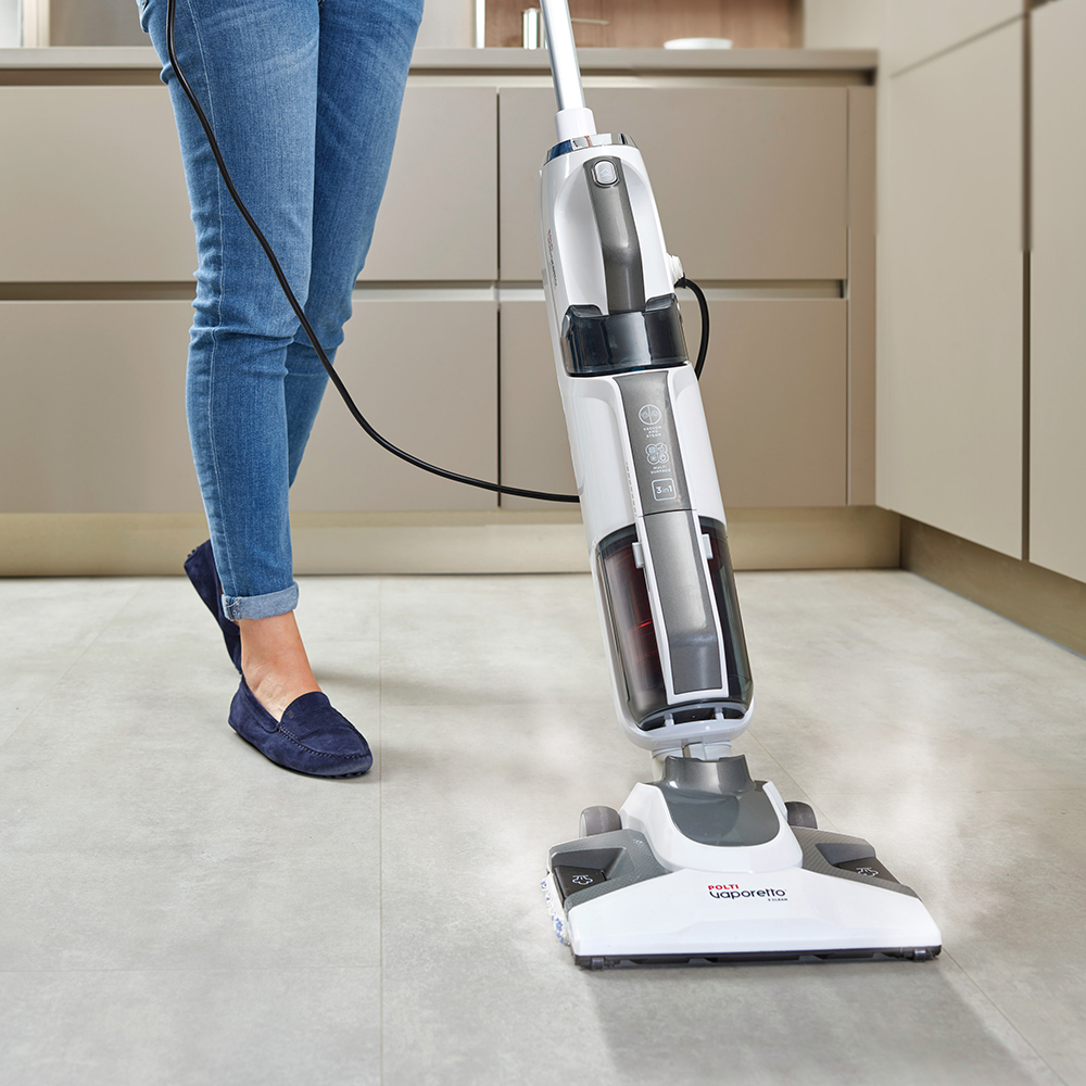 Una persona usa una scopa a vapore bianca e grigia su un pavimento piastrellato. Indossa jeans e scarpe. I mobili della cucina sono sullo sfondo.