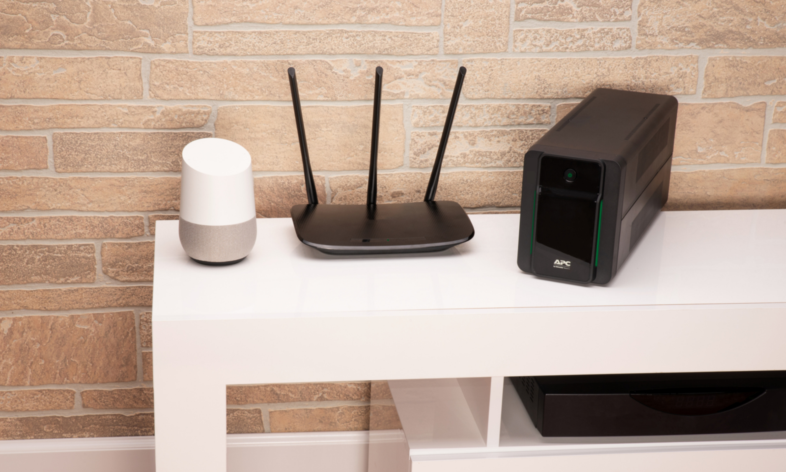 Biały i szary głośnik, czarny router z antenami i czarny zasilacz UPS APC na białej półce na tle ceglanej ściany.