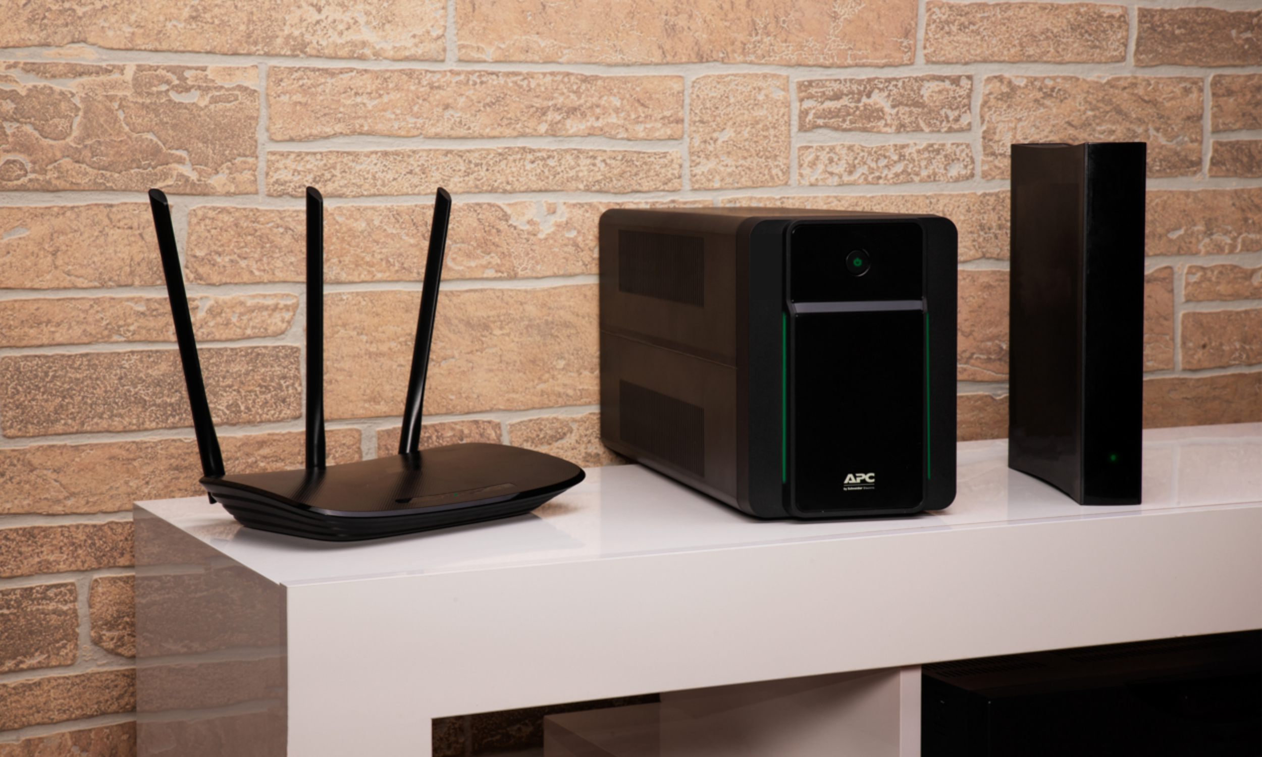 Schwarzer Router, APC USV und ein schwarzer Server stehen auf einem weißen Regal vor einer Backsteinmauer.