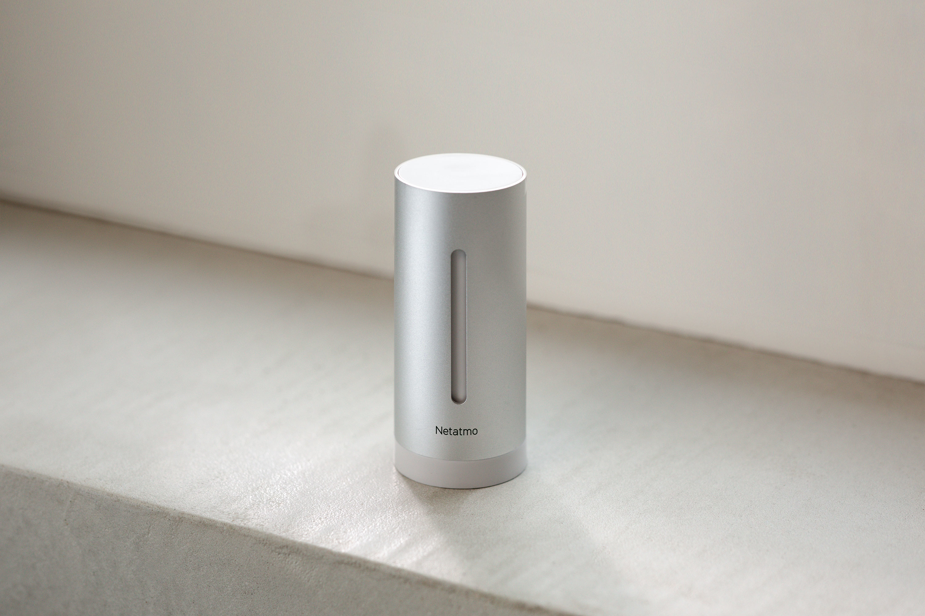 A silver Netatmo weather station stands on a light surface. The device is cylindrical with a vertical slot.