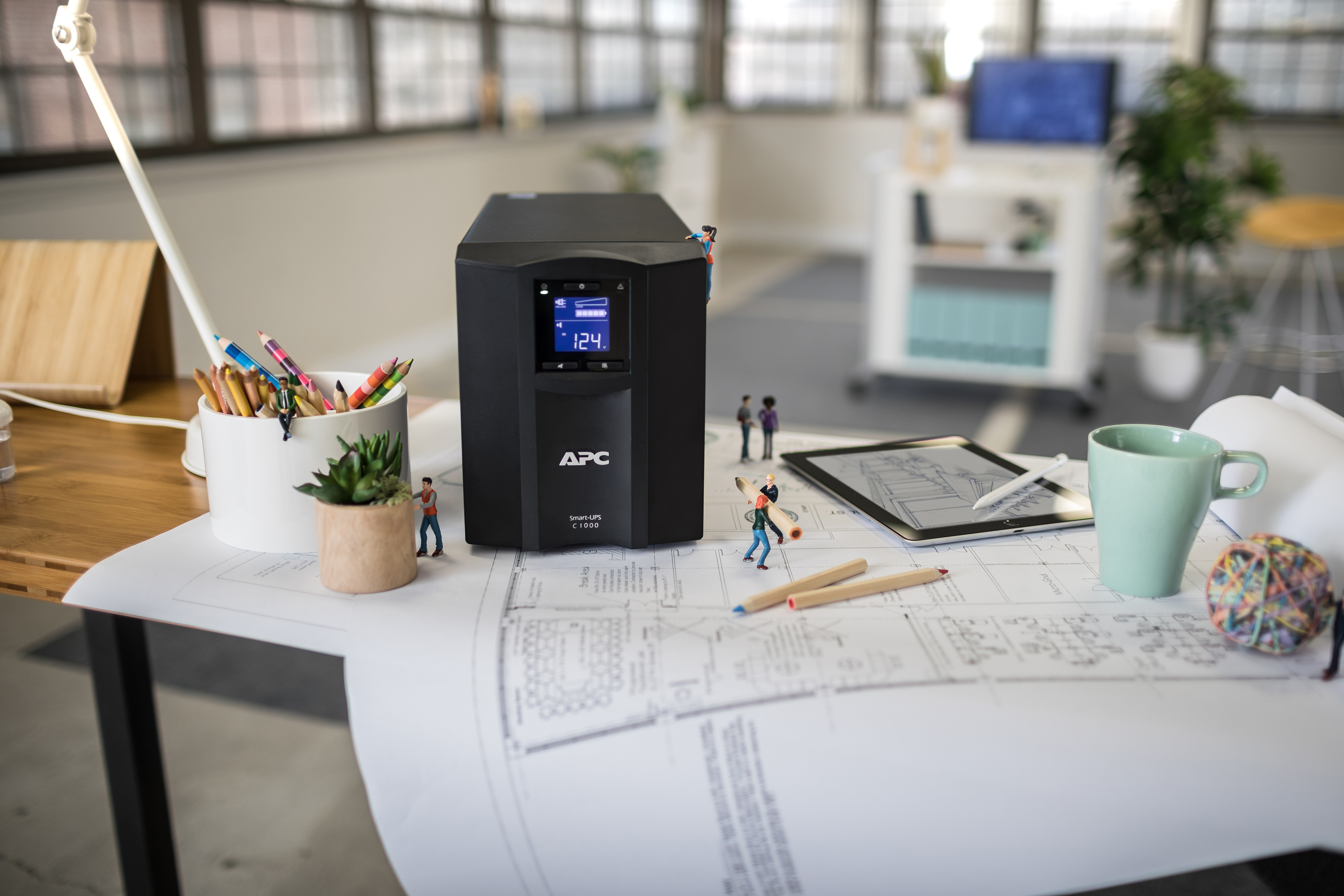 Ein Schreibtisch mit einer USV von APC, Bleistiften, einer Tasse und einem Tablet. Miniaturfiguren sind um die Objekte platziert.