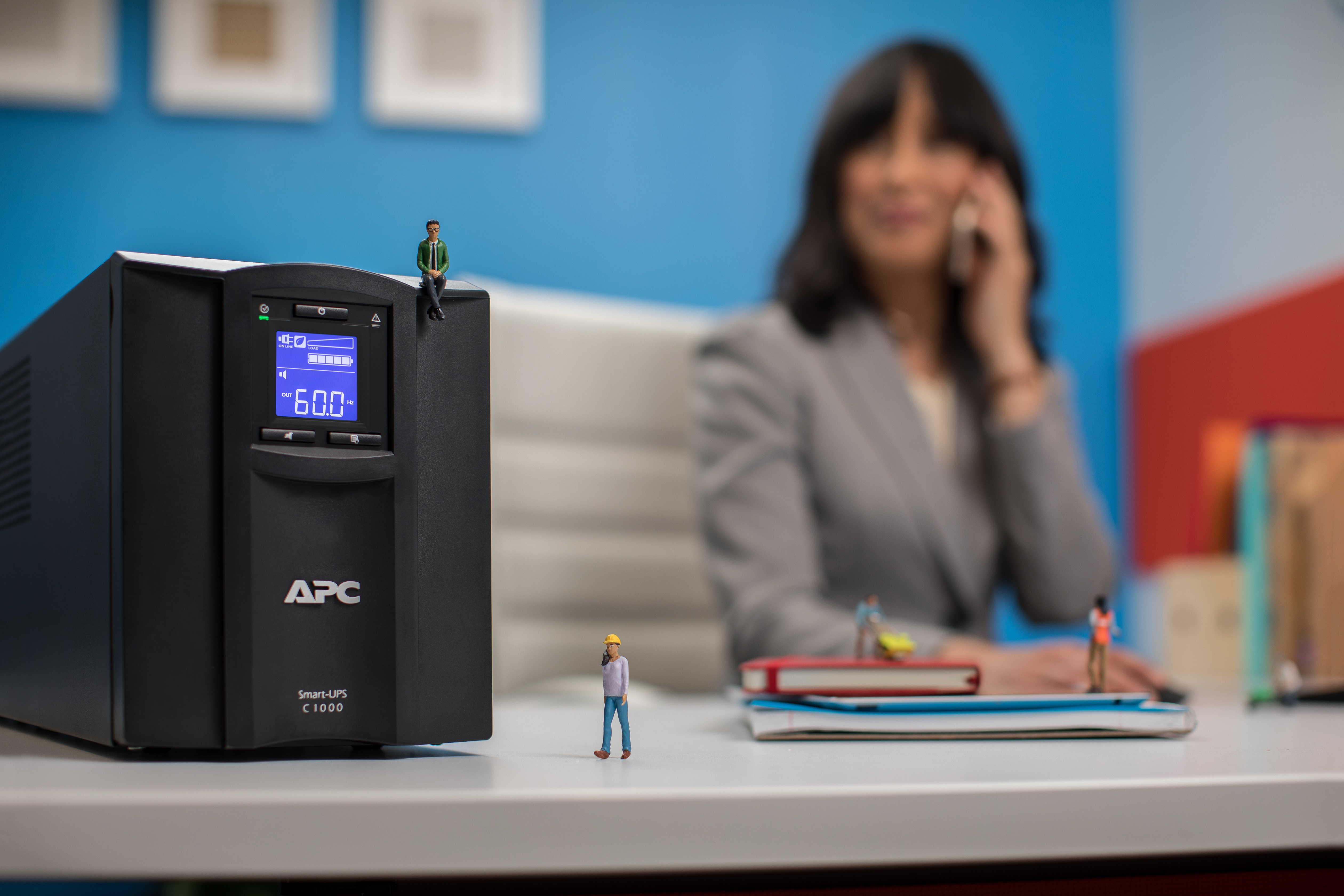 Eine USV von APC mit Miniaturfiguren. Im Hintergrund telefoniert eine Frau. Andere Figuren befinden sich in der Nähe der USV.