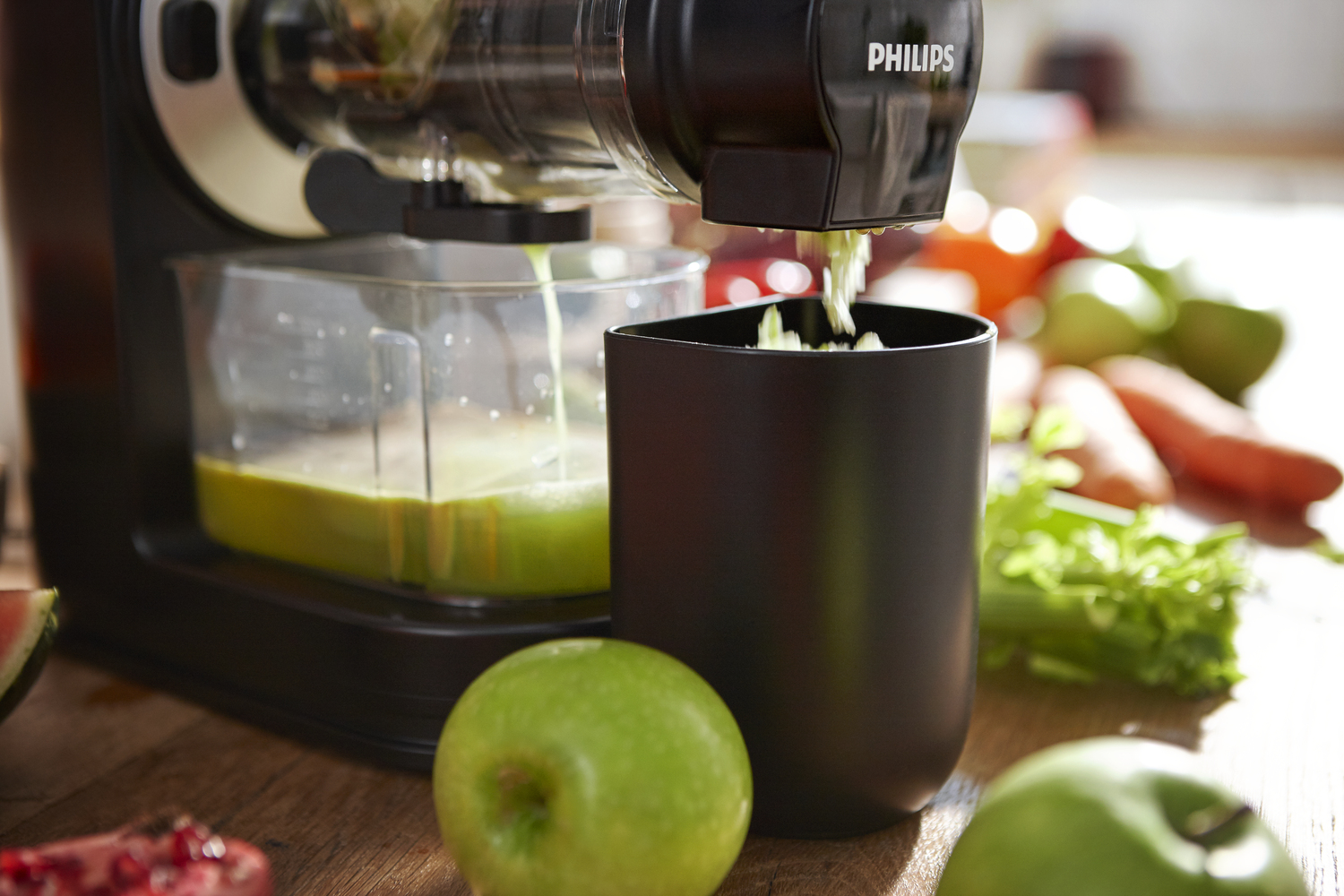 Ein Entsafter extrahiert Saft. Ein schwarzer Behälter füllt sich mit Fruchtfleisch. Grüner Apfel im Vordergrund, Gemüse im Hintergrund.