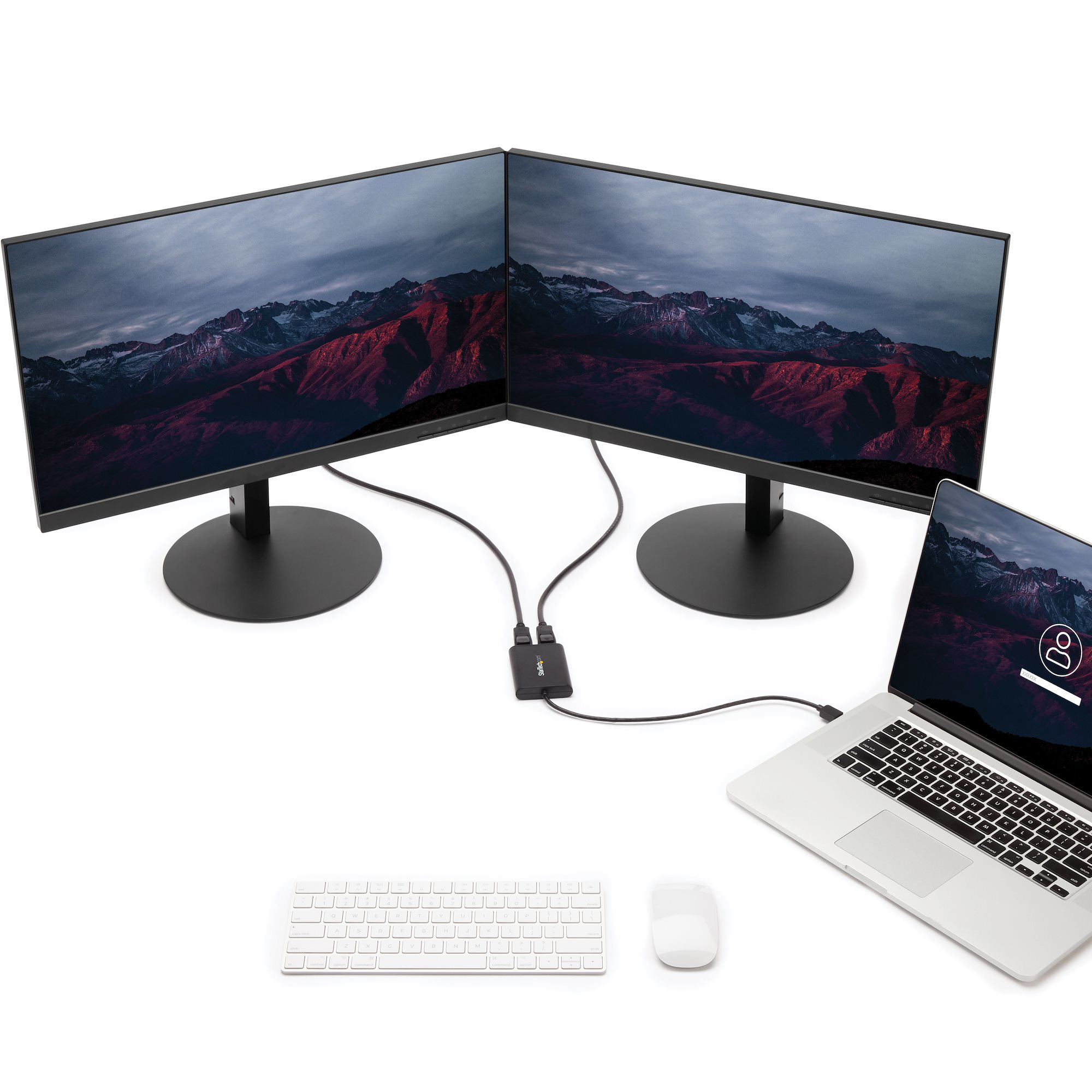 Dos monitores con escena montañosa, conectados a laptop y hub, teclado y ratón sobre superficie blanca.