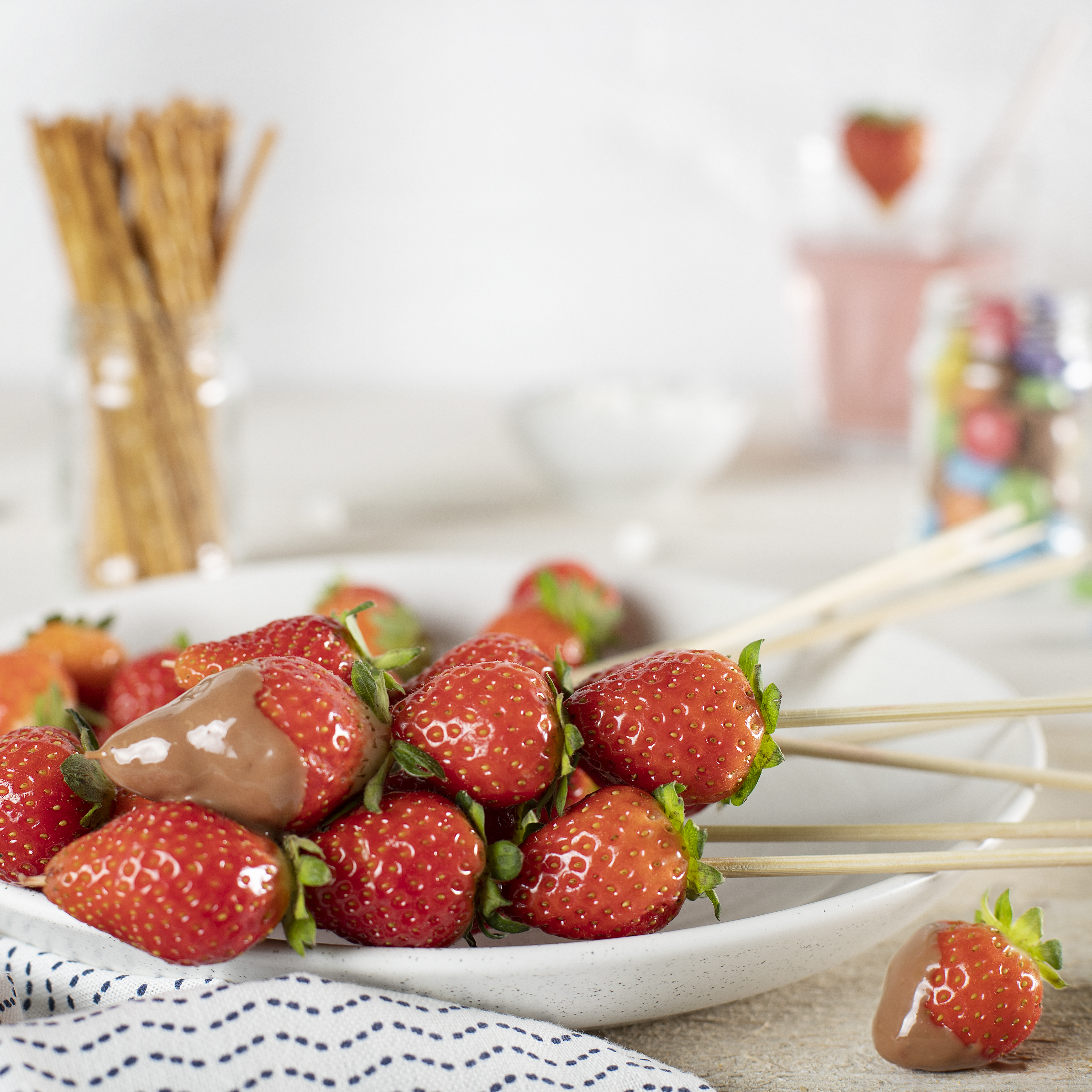 Erdbeeren auf Spießen, einige in Schokolade getaucht, auf einem weißen Teller. Im Hintergrund Snacks und Zutaten.
