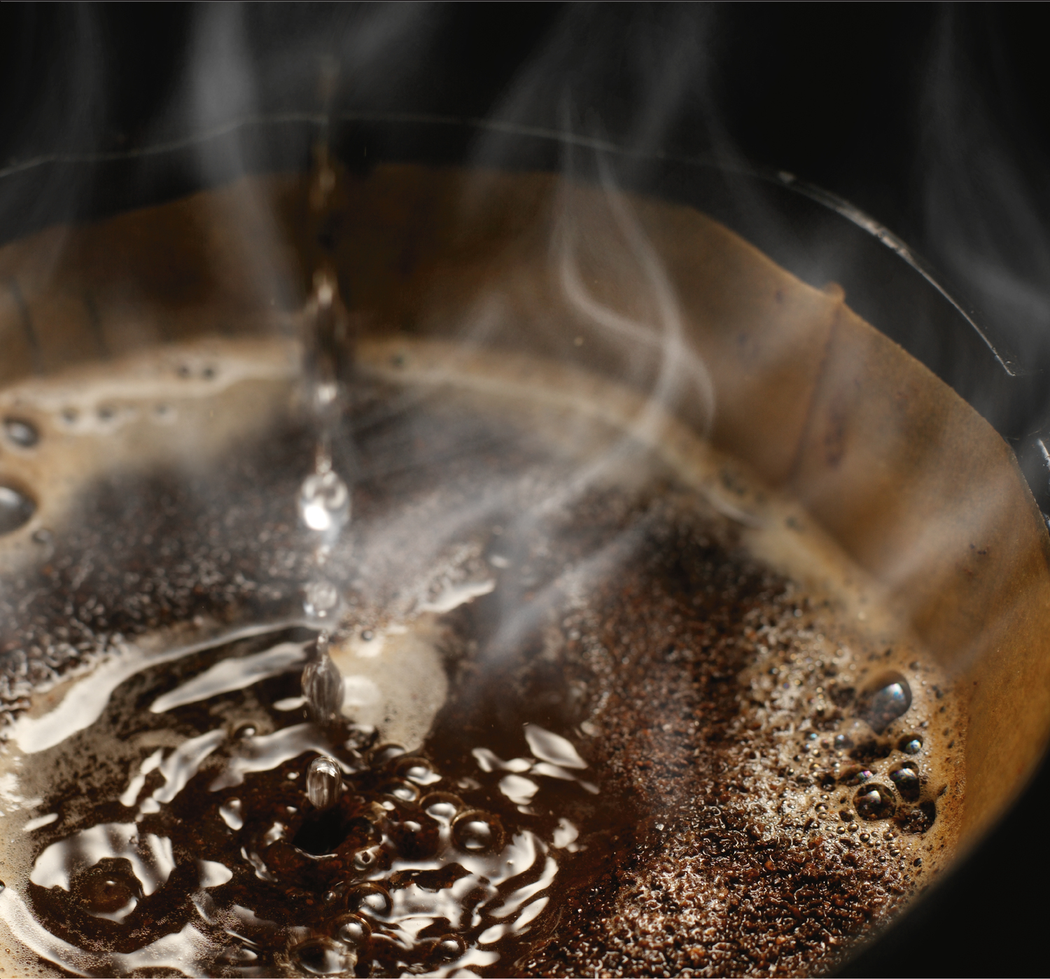 Zbliżenie parzenia kawy. Woda kapie do brązowego filtra z fusami kawy, tworząc bąbelki. Para unosi się na czarnym tle.