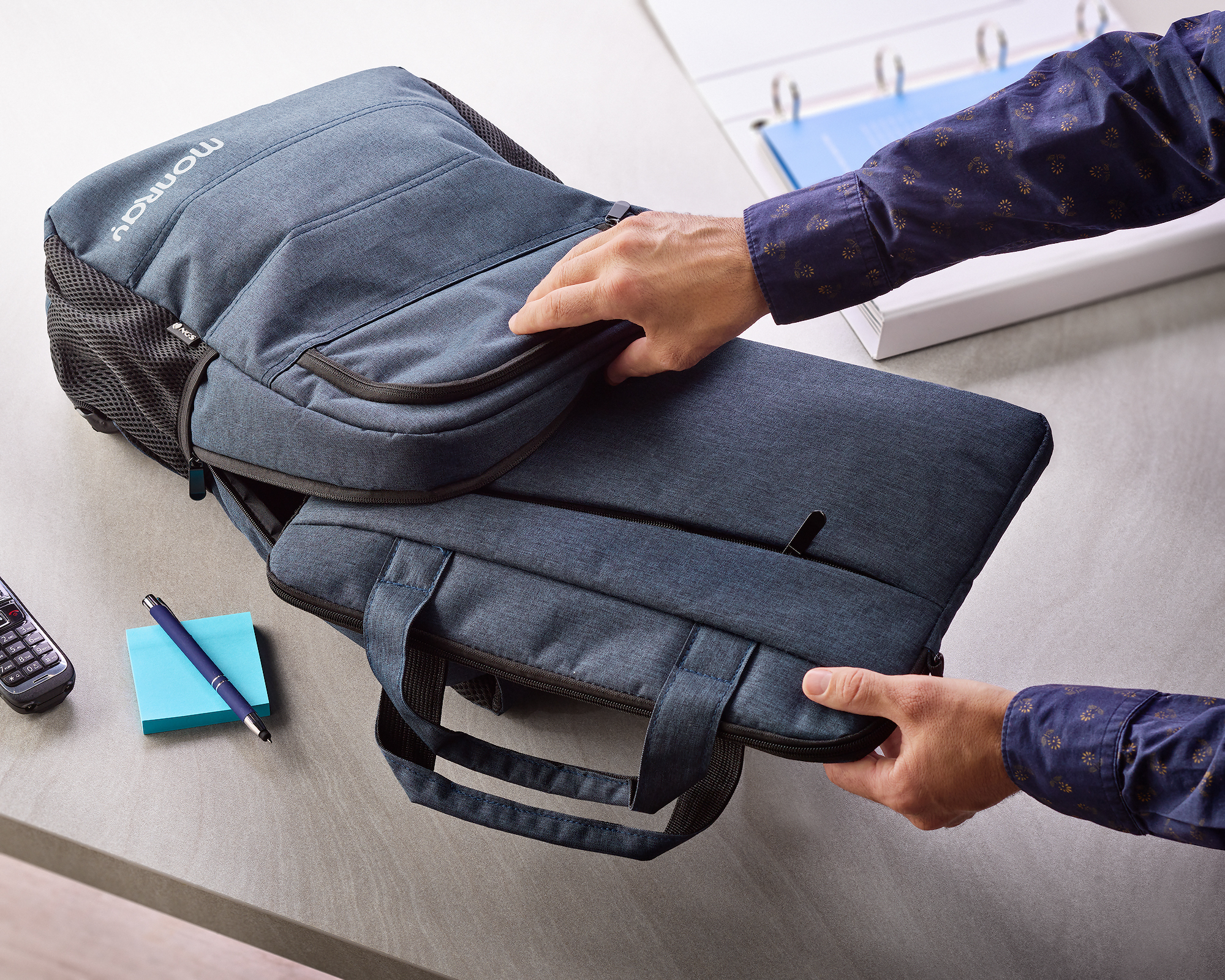 Una persona sta aprendo una borsa blu per laptop. Una penna blu, post-it e un telefono sono sul tavolo.