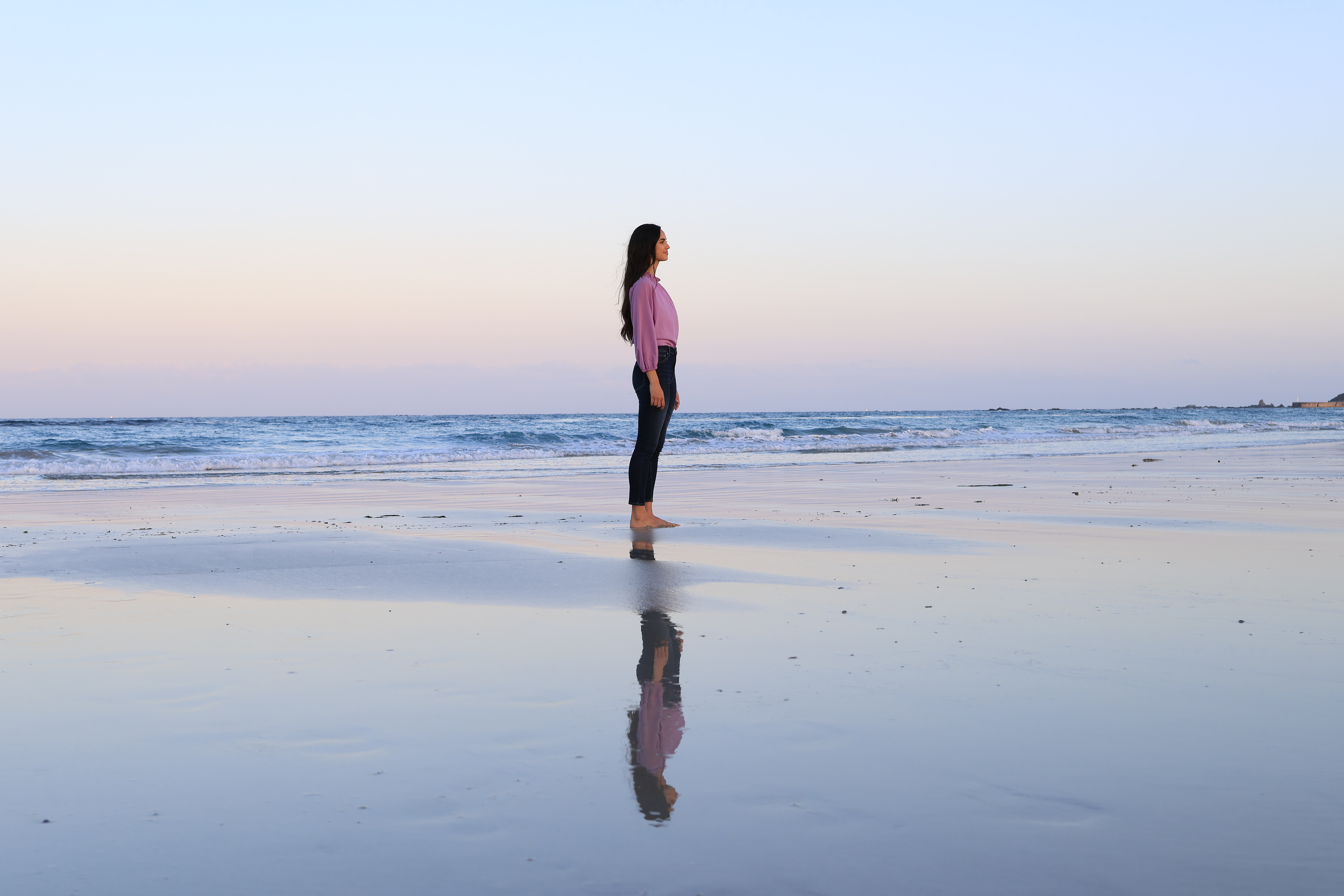 Eine Frau steht barfuß am nassen Strand, ihr Spiegelbild sichtbar. Der Himmel ist pastellfarben, das Meer ruhig.