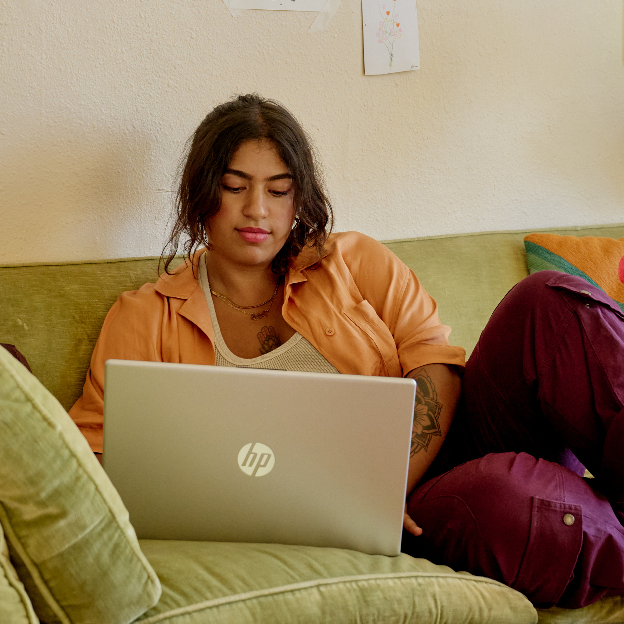 Mujer en un sofá verde usando una laptop HP. Lleva camisa naranja y pantalones morados. El fondo es una pared beige.