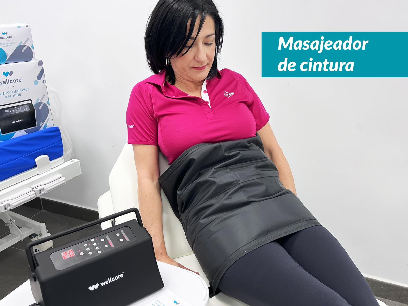 Mujer con un masajeador de cintura. Lleva una camisa rosa y pantalones negros. Hay una máquina a la izquierda.