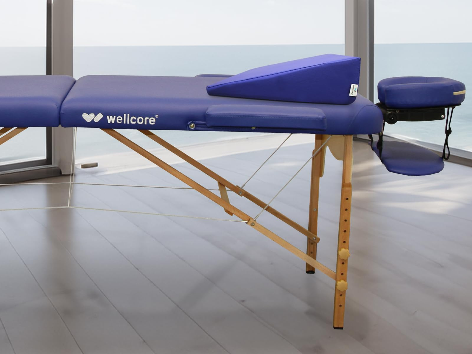 Una camilla azul con reposacabezas y almohada. La mesa es de madera y el fondo es una ventana con vista al mar.