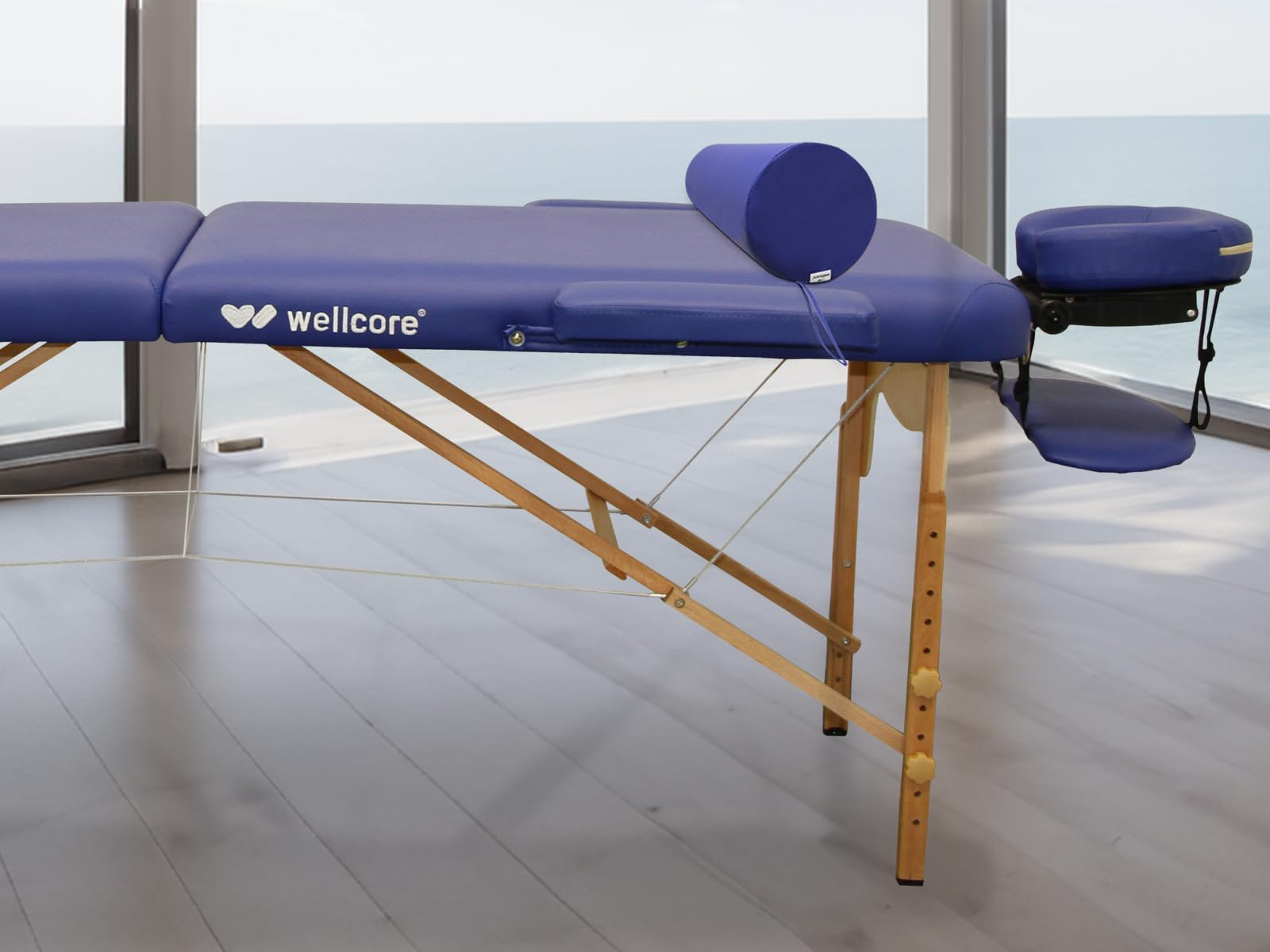 Una mesa de masaje azul junto a una ventana con vista al mar. Tiene reposacabezas, reposabrazos y un cojín cilíndrico.