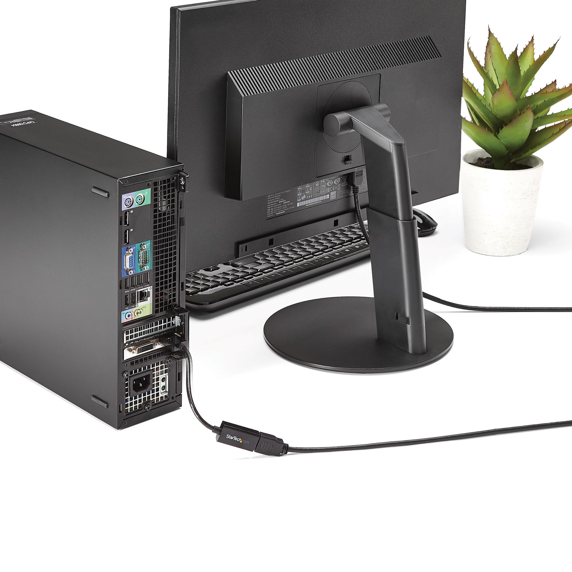 Una configuración de computadora con monitor negro, torre de PC, teclado y planta. Cables conectan dispositivos.