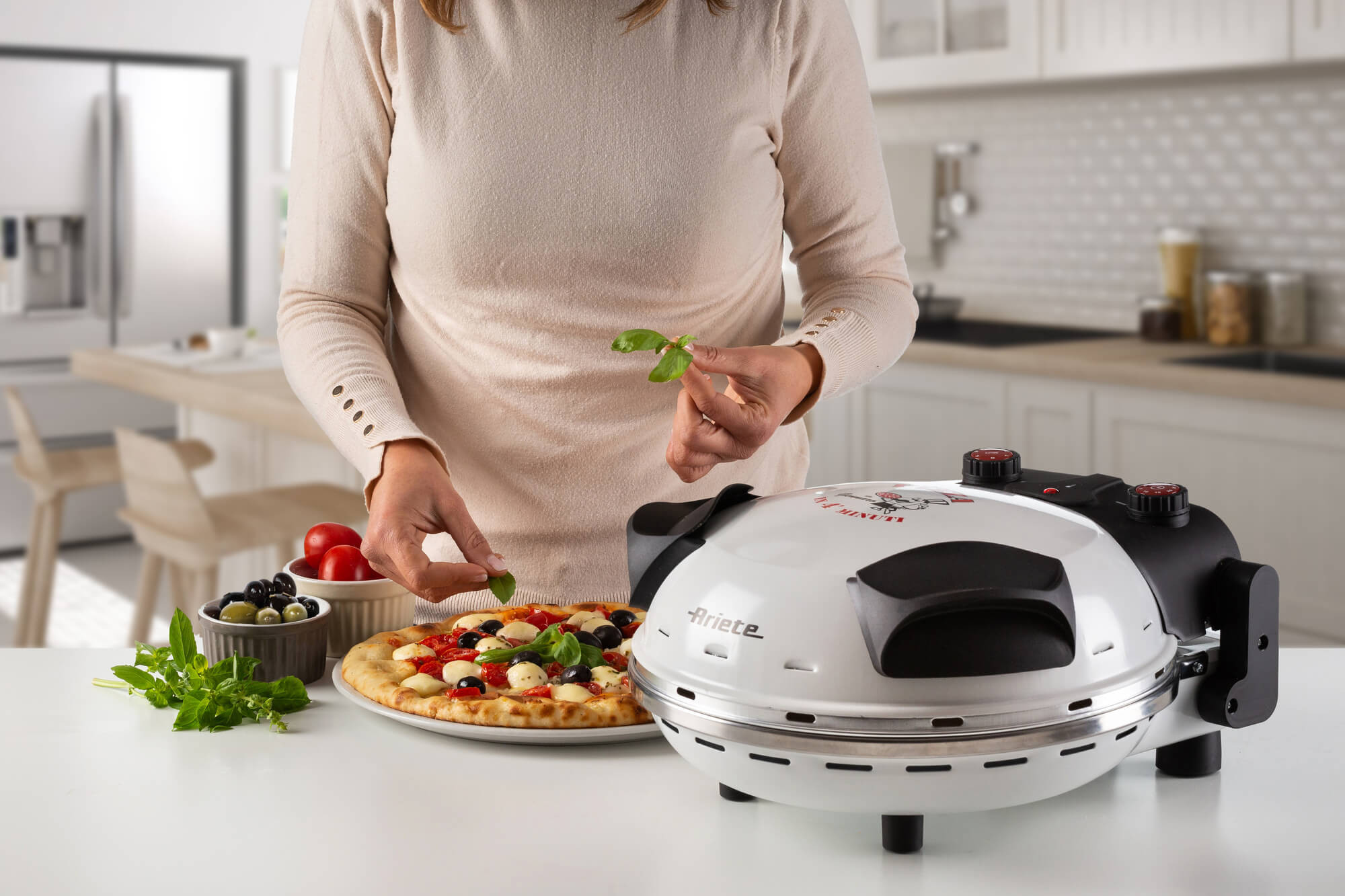 Eine Person belegt Pizza mit Basilikum, neben einem Pizzaofen. Zutaten und Pizza stehen auf einem weißen Tisch in einer Küche.