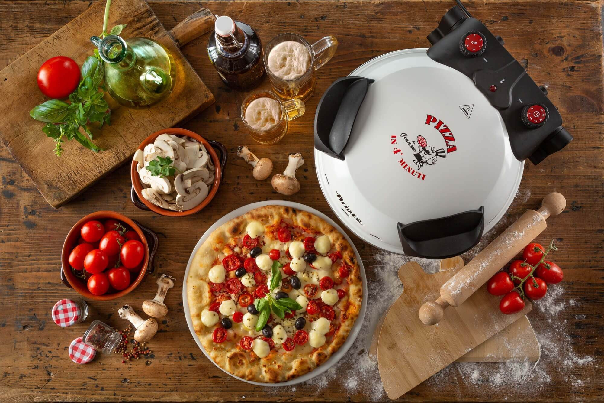 Pizza mit Tomaten, Mozzarella und Oliven, mit Zutaten und Pizzaofen auf einem Holztisch. Bier und Pilze sind vorhanden.