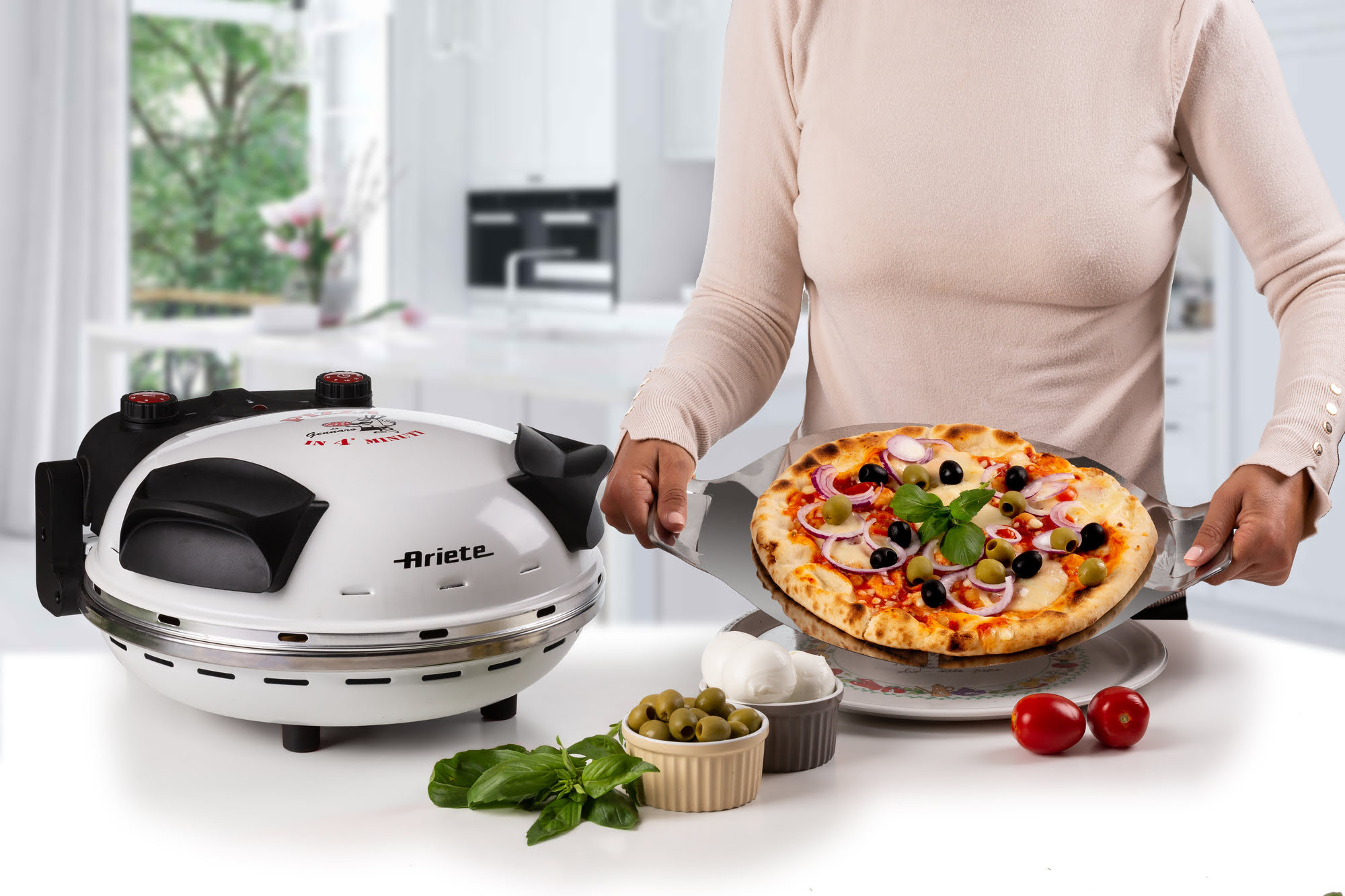 Eine Person hält eine Pizza mit Belag. Ein weißer Pizzaofen und Zutaten wie Oliven und Tomaten stehen auf dem Tisch.
