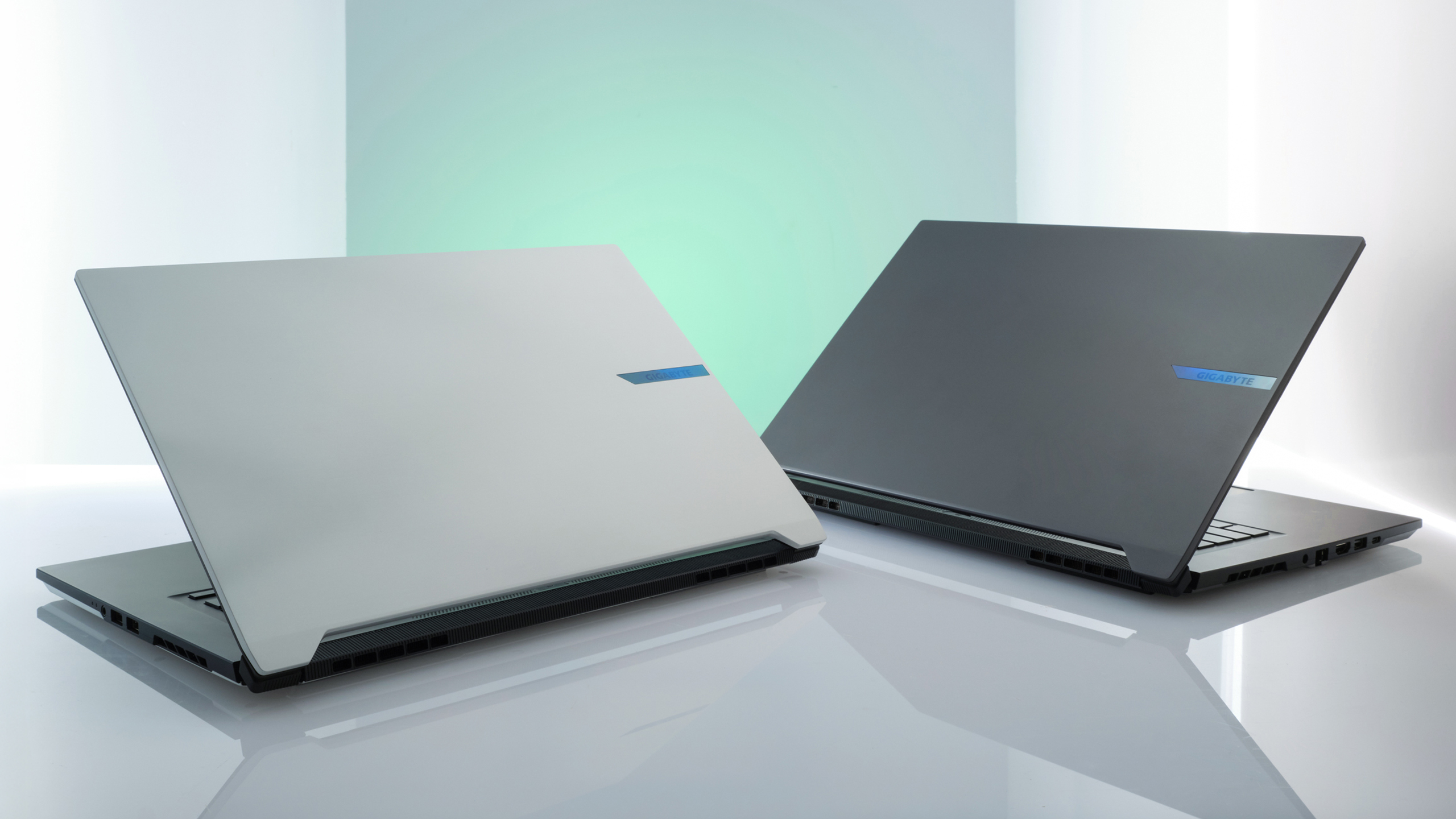 Dwa otwarte laptopy na odblaskowej powierzchni. Jeden biały, drugi czarny, oba z niebieskimi akcentami. Tło jest jasne i ma gradienty.