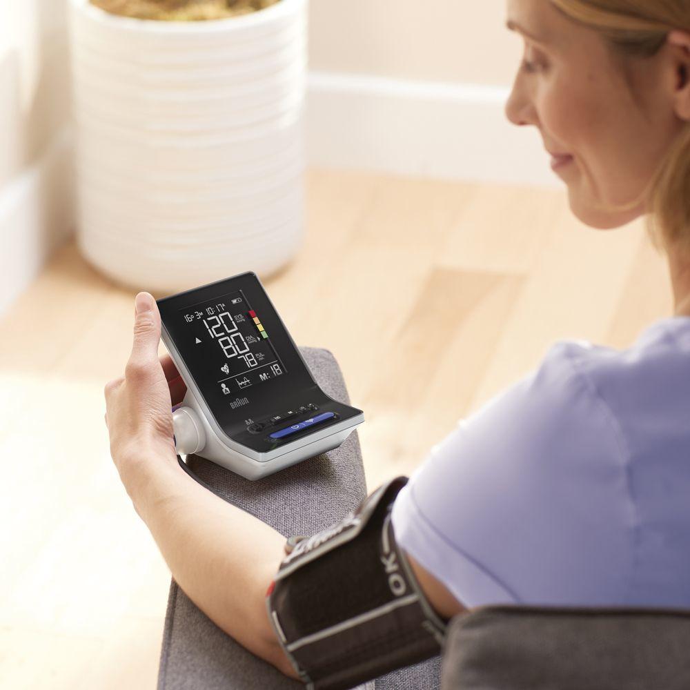 Una mujer controla la presión arterial con un monitor. El monitor muestra números. Un brazalete está en su brazo. Una planta está en el fondo.